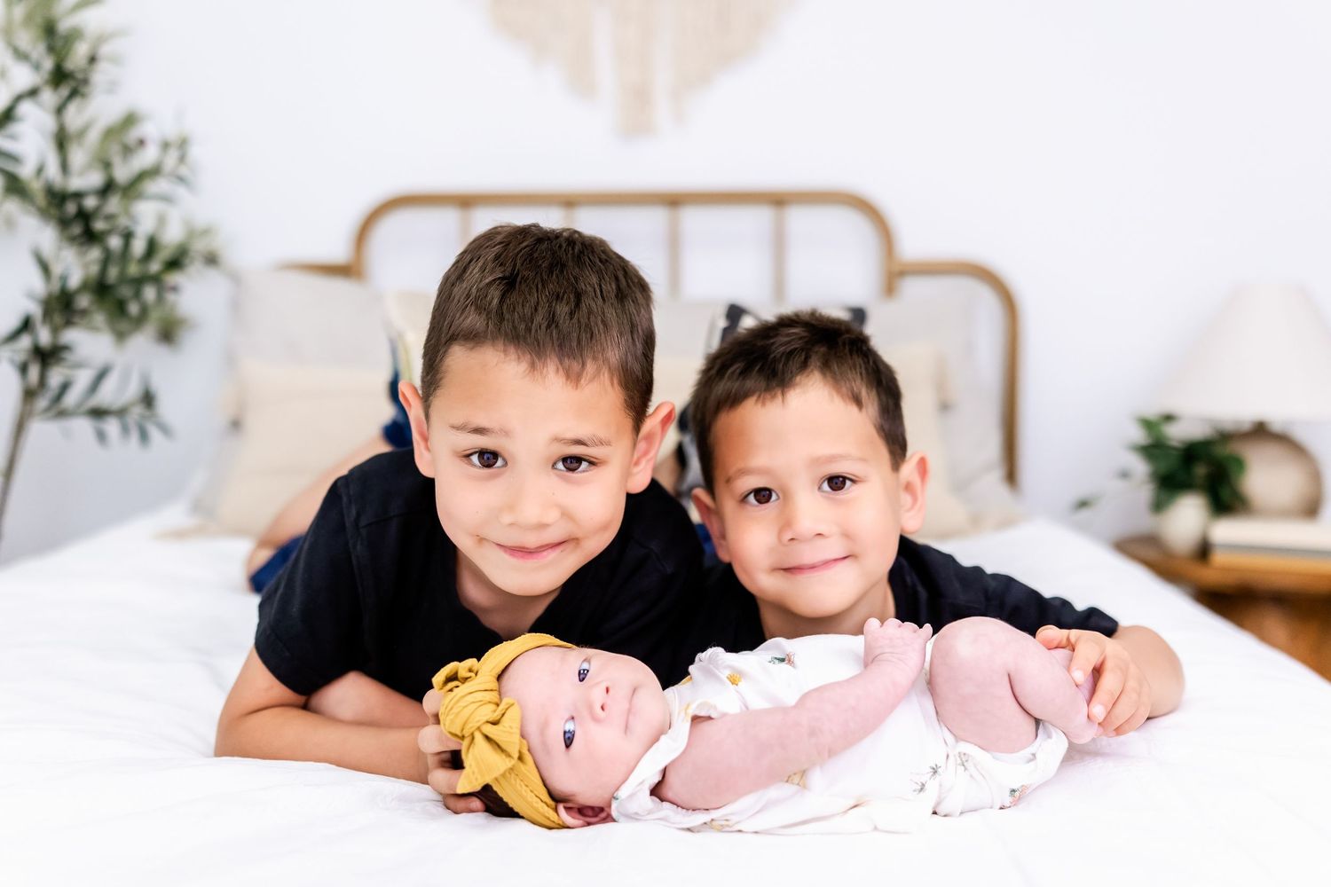 Two young siblings lie together on a white bed with baby dolls in a bright bedroom setting.