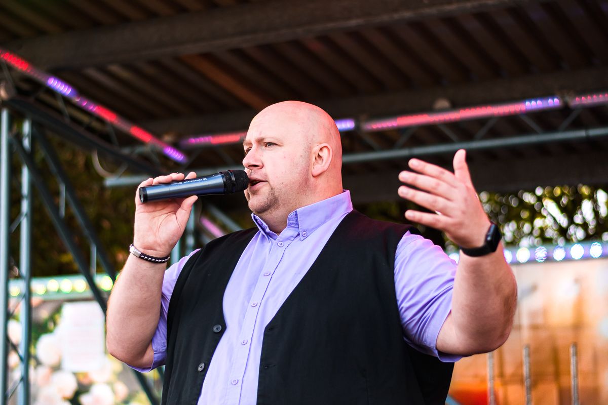 Singer in purple shirt and black vest performs passionately on outdoor stage with expressive hand gesture.