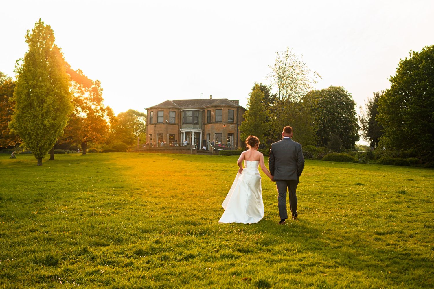 A couple walks away from a grand house at sunset across a grassy lawn.