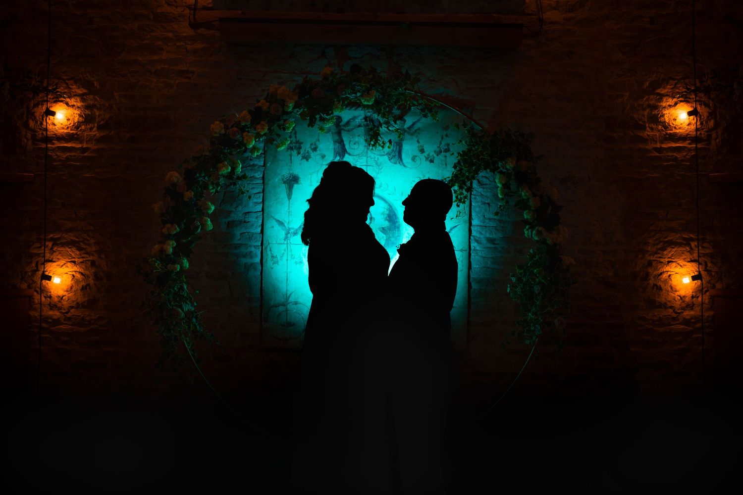 Silhouettes of two figures stand in a stone archway illuminated by warm lanterns and ethereal turquoise backlighting.