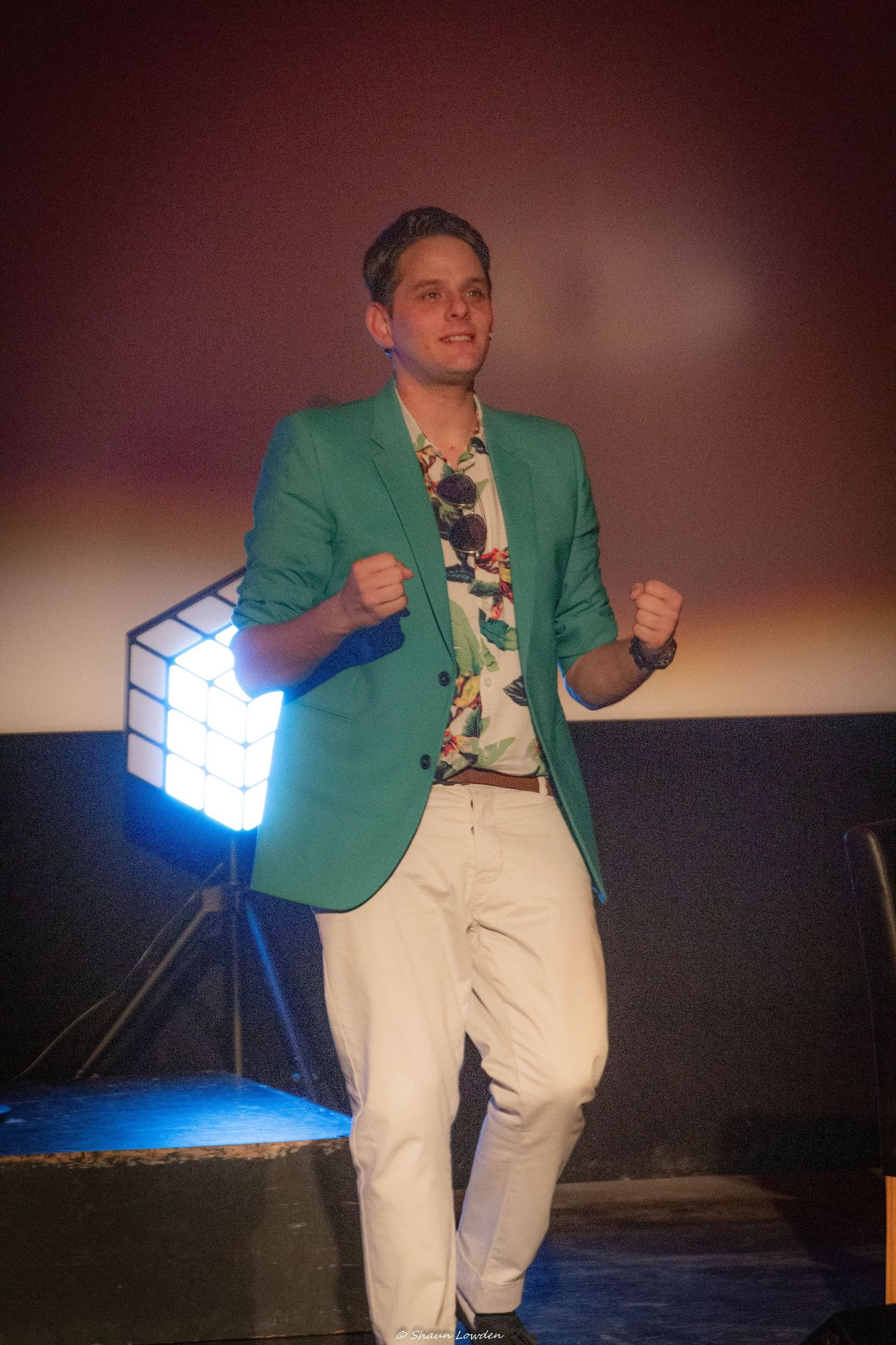 Stage performer in mint green blazer and white pants gestures energetically near a stage light.