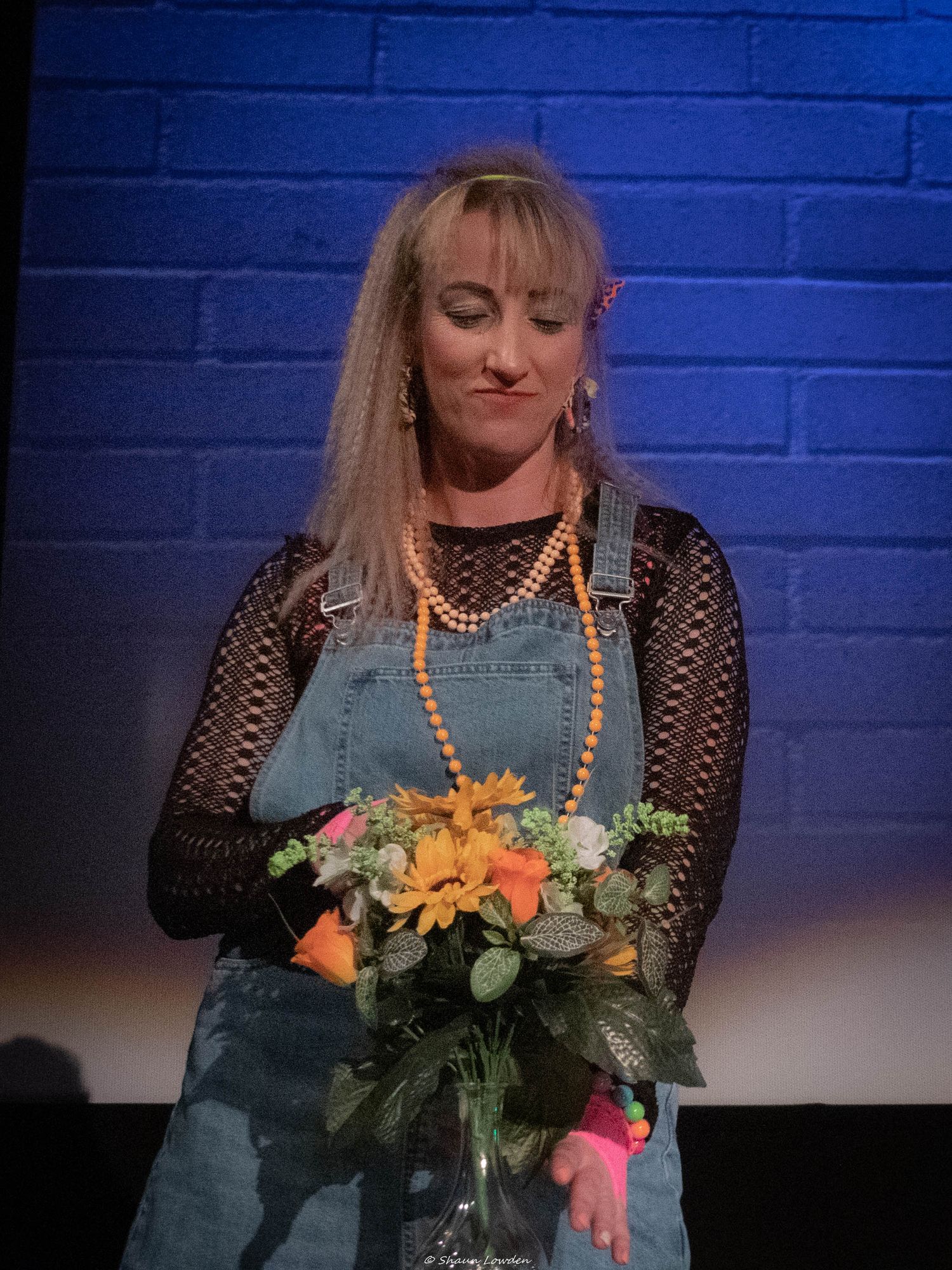 Stage performer in denim overalls holds flower bouquet against blue brick wall backdrop.
