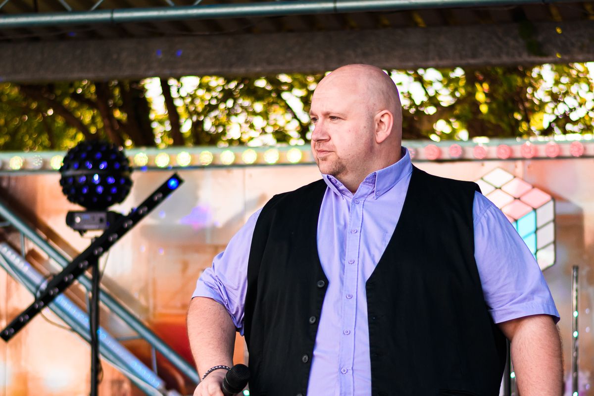 A performer in a black vest and purple shirt stands at a microphone on an outdoor stage during a summer event.