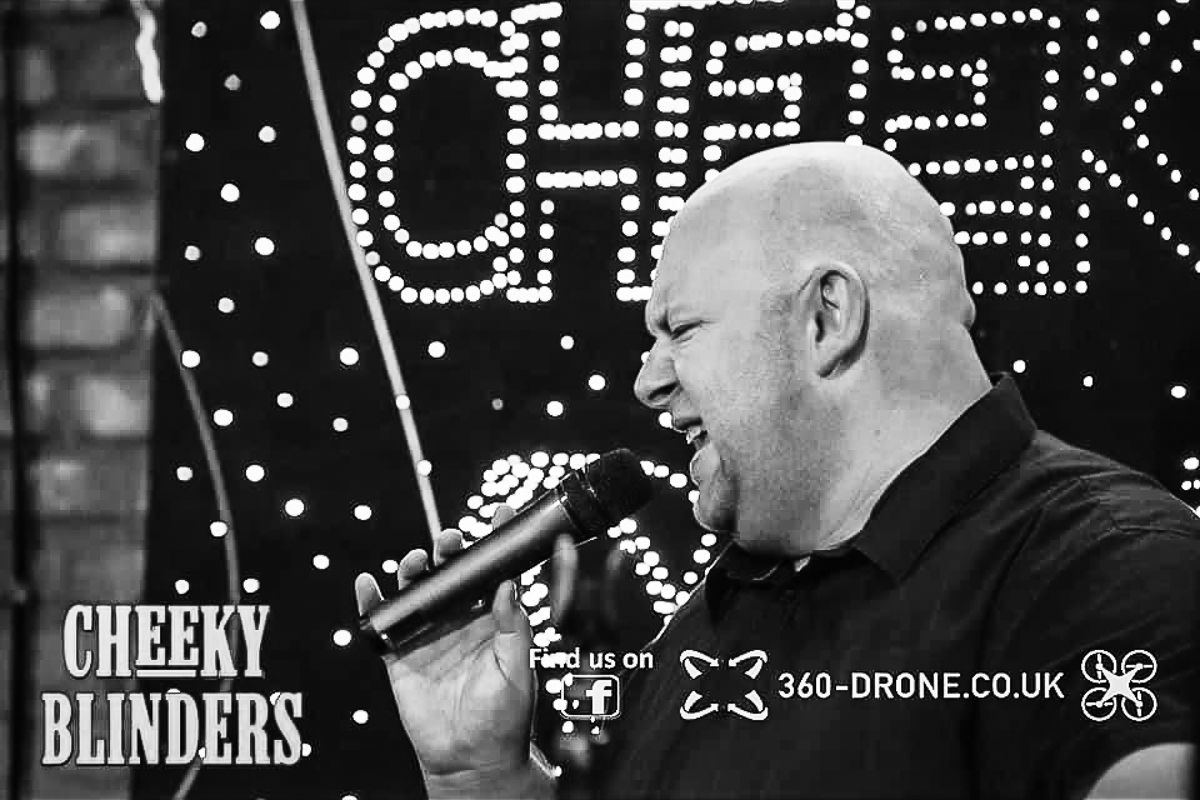 Performer singing passionately against illuminated 'Cheeky' sign backdrop in black and white photo.