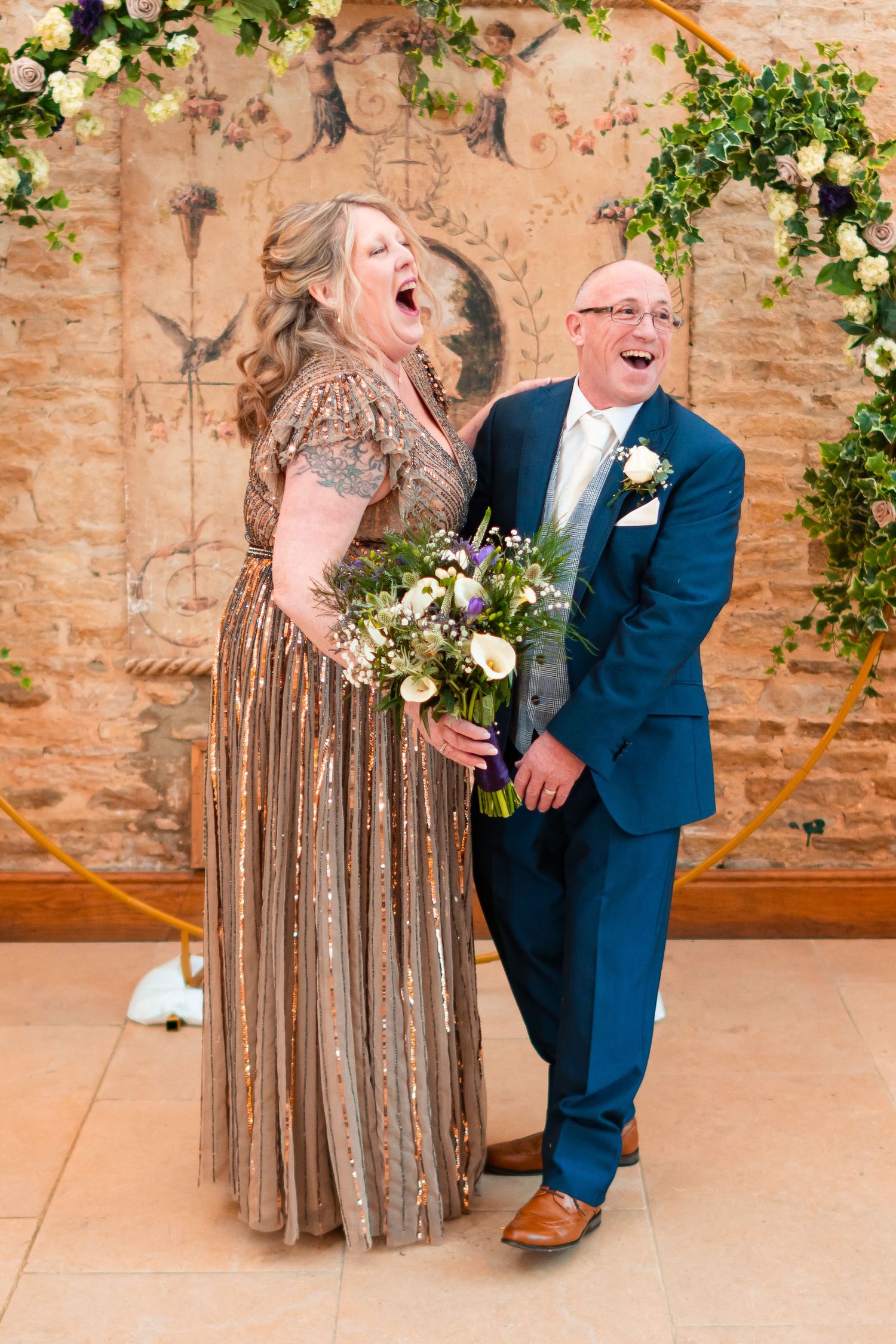 A couple shares a joyful laugh together at their wedding, wearing a sparkly gold gown and navy blue suit.