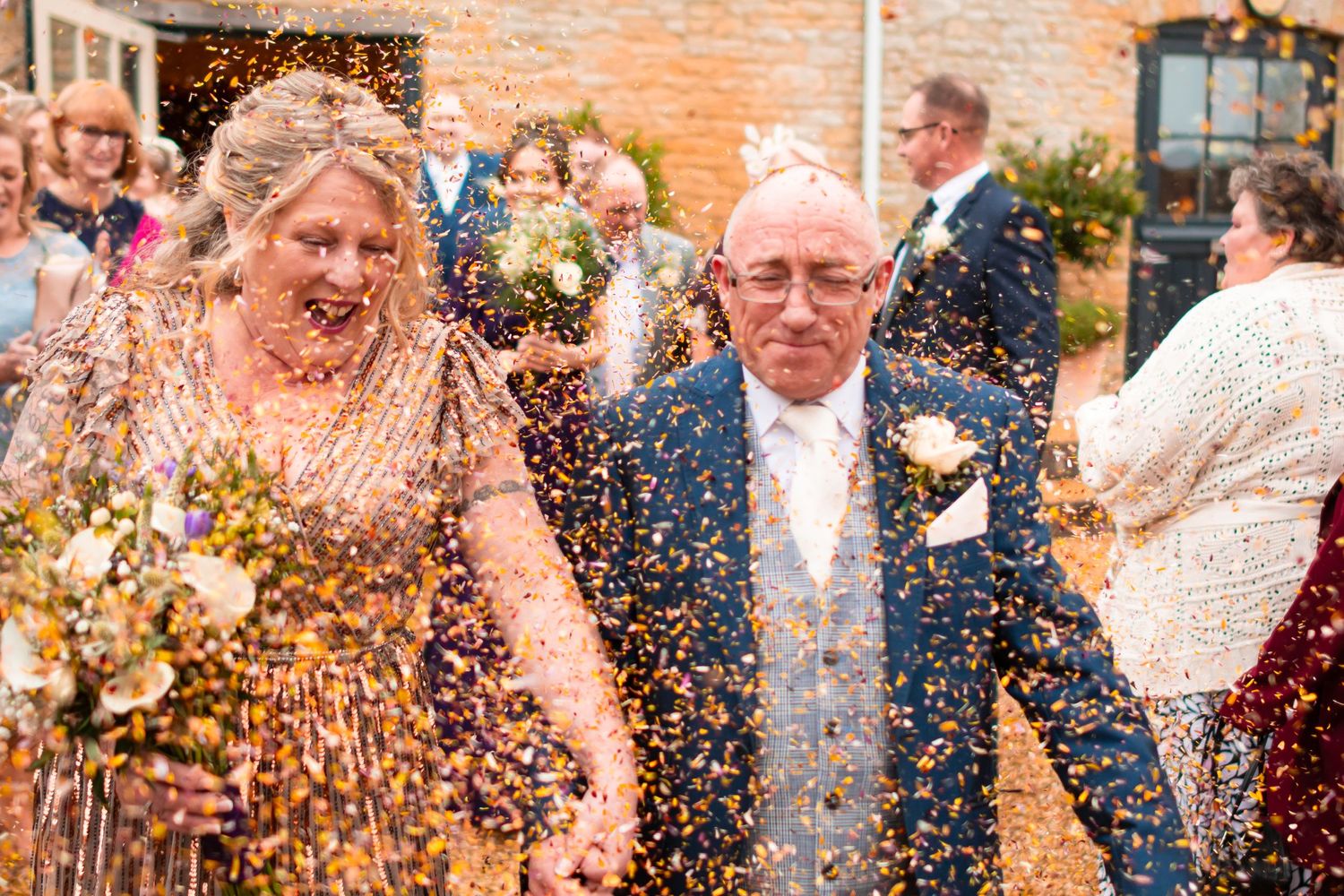Joyful wedding celebration moment with confetti being thrown at a brick wall venue in warm natural lighting.