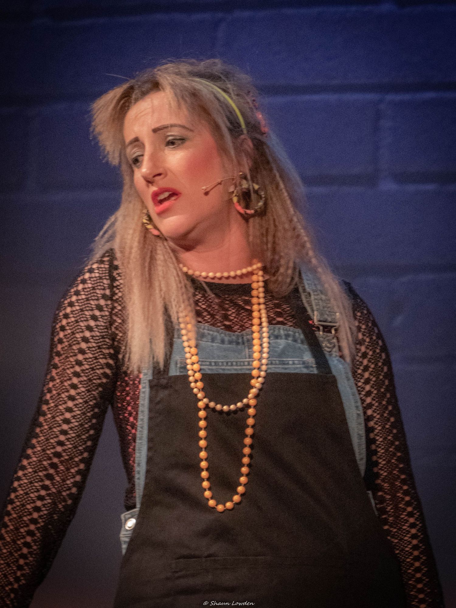 Performer in mesh top and overalls with orange beads against blue backdrop during stage show.