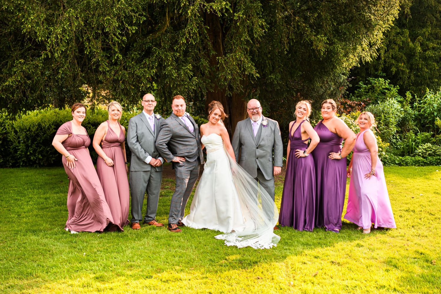 Wedding party poses together on a sunny lawn with bridesmaids in pink and purple dresses.
