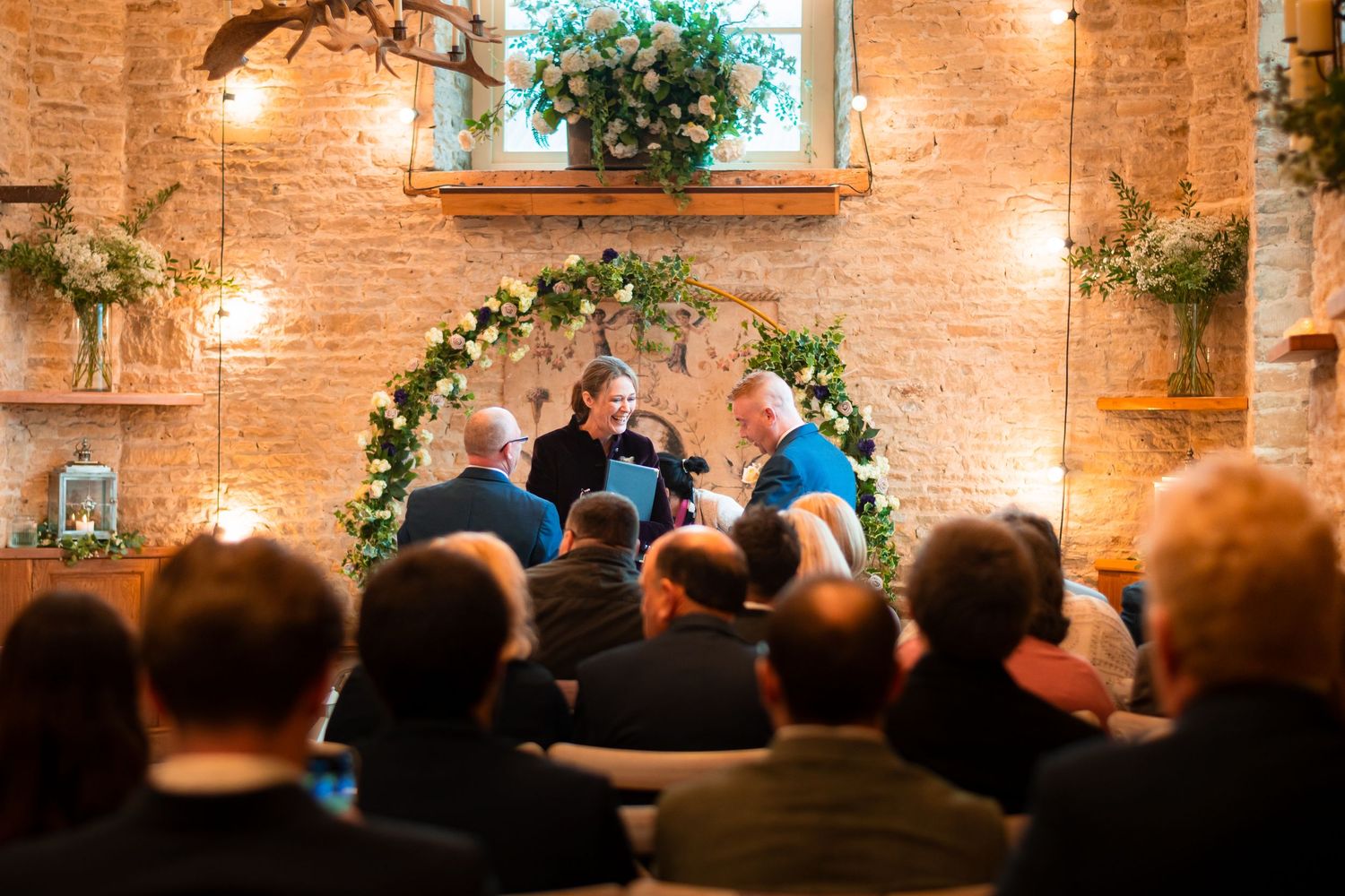 A small ceremony takes place in a rustic brick venue adorned with greenery and warm glowing lights.