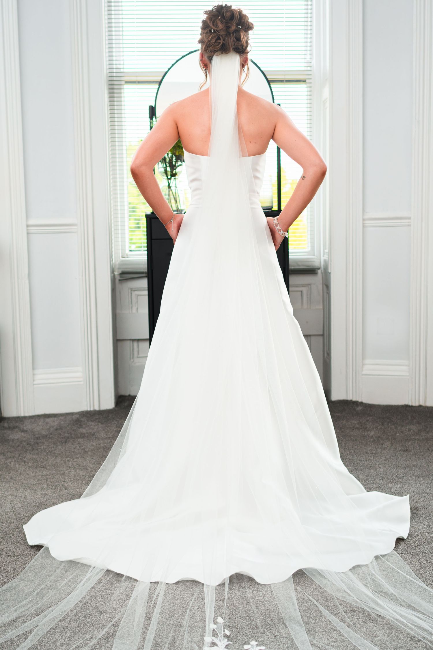 Back view of a wedding dress with flowing white train standing against white window backdrop.