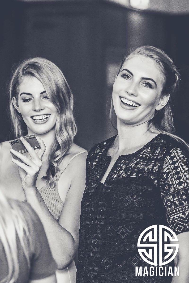 Two women smile joyfully at a nightclub with Magician logo, one in beige and one in black patterned tops.