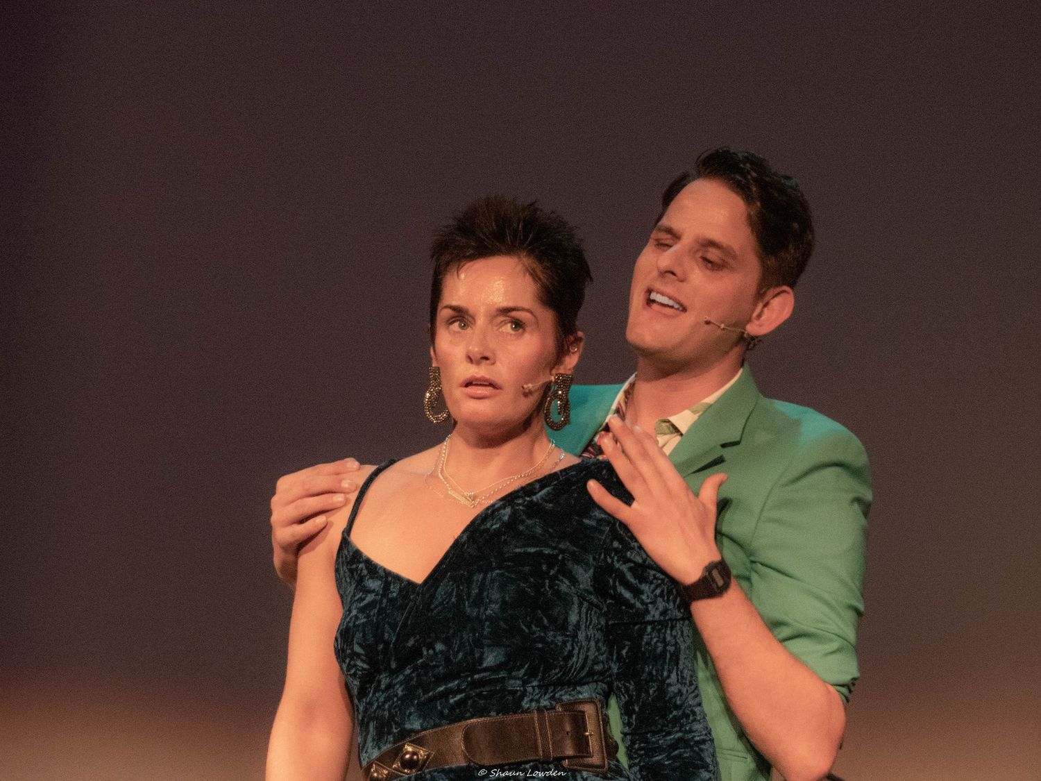 Two performers in dramatic embrace during theatrical scene with green blazer and velvet dress.