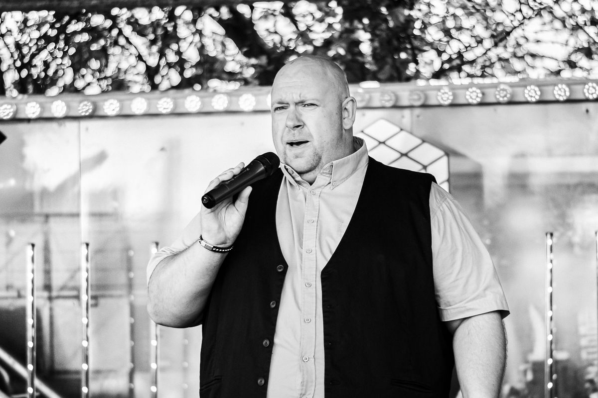 Black and white photo of performer singing emotionally with microphone on outdoor stage.
