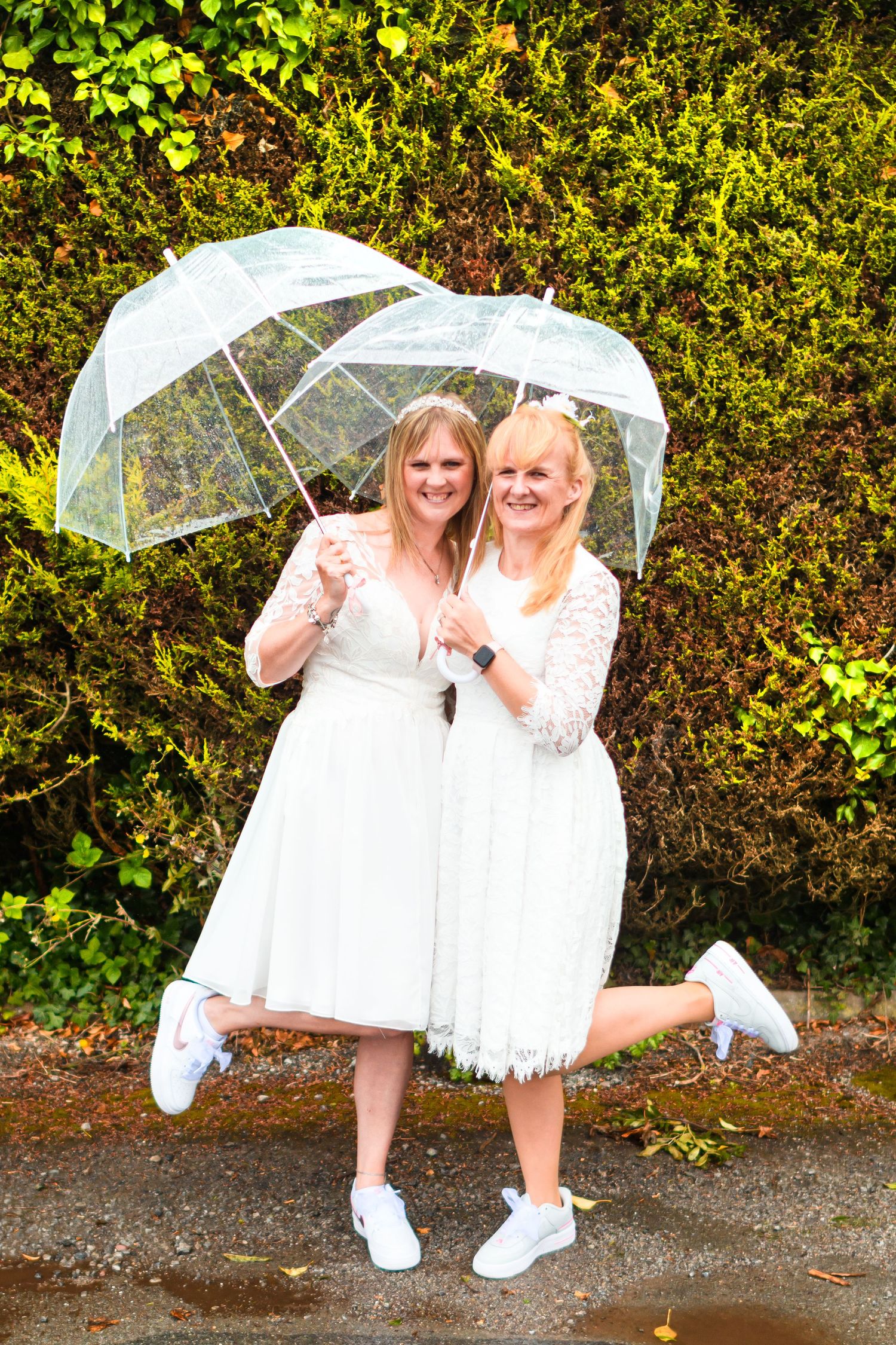 Two people in white dresses pose playfully with clear umbrellas in a garden setting.