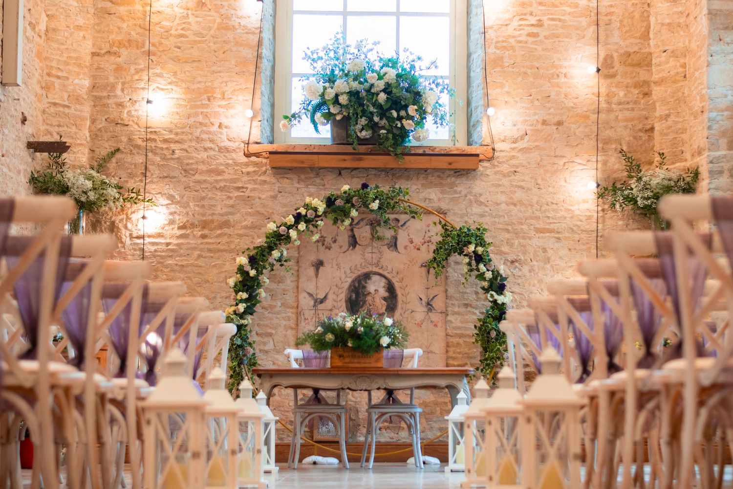 Rustic wedding ceremony setup with wooden chairs and floral arch in exposed brick venue with window light.
