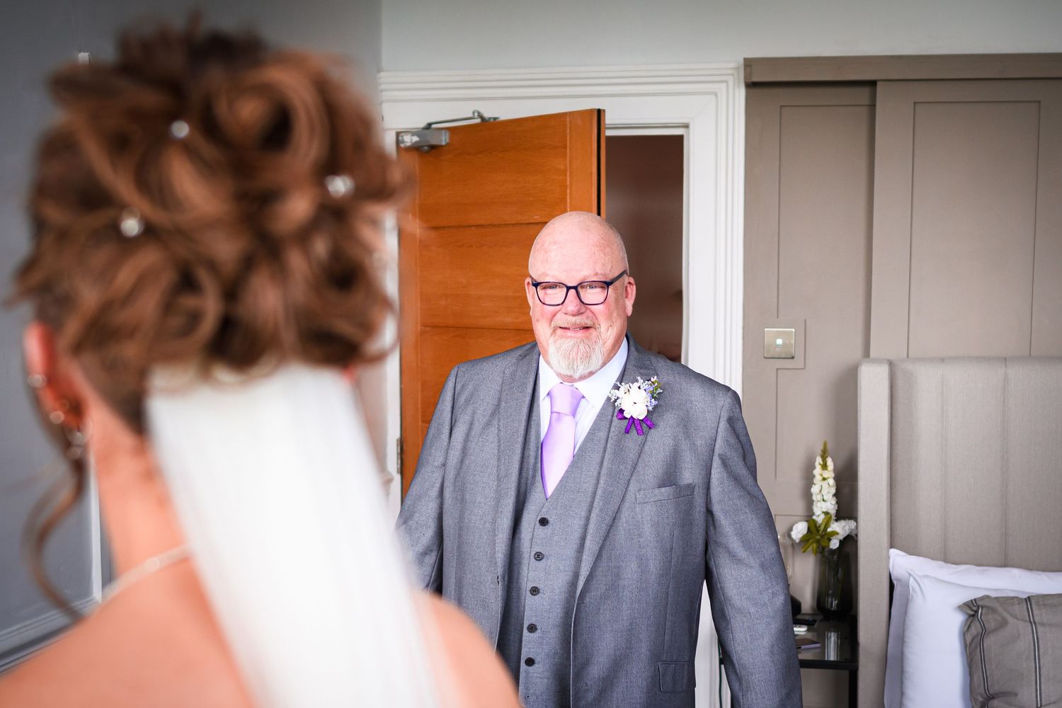 Person in grey suit with boutonniere standing in doorway during wedding preparation.