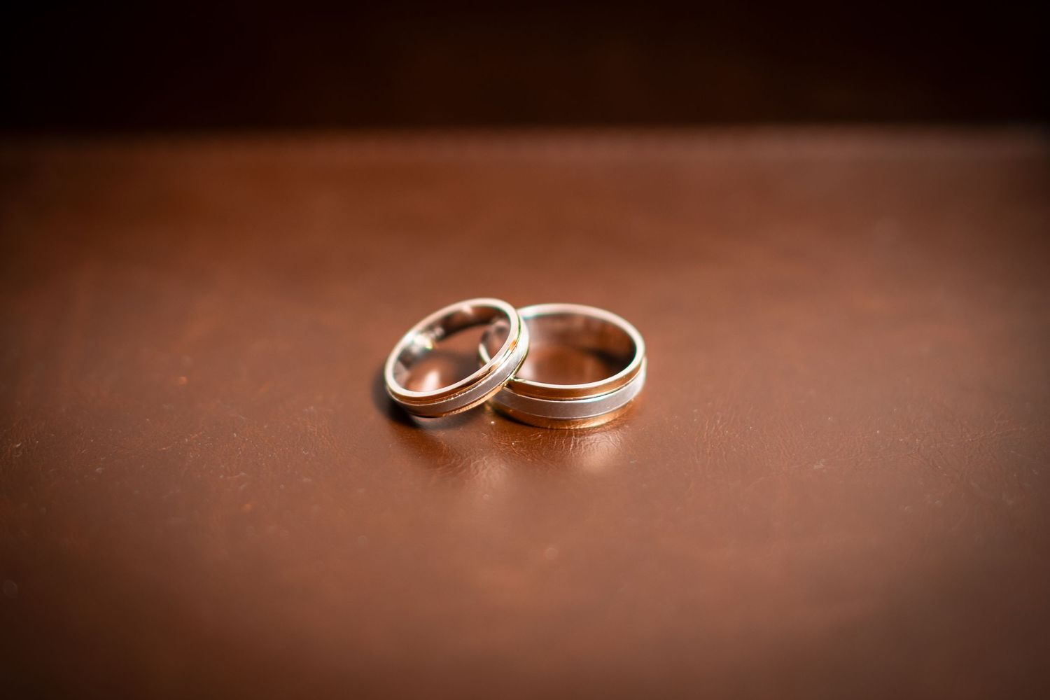 Two elegant wedding bands with metallic finishes rest together on a brown leather surface in soft lighting.