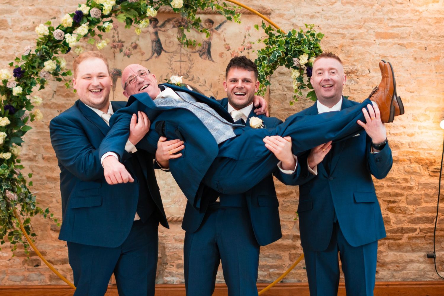 Four people wearing matching navy blue suits pose playfully against a brick wall decorated with green vines.