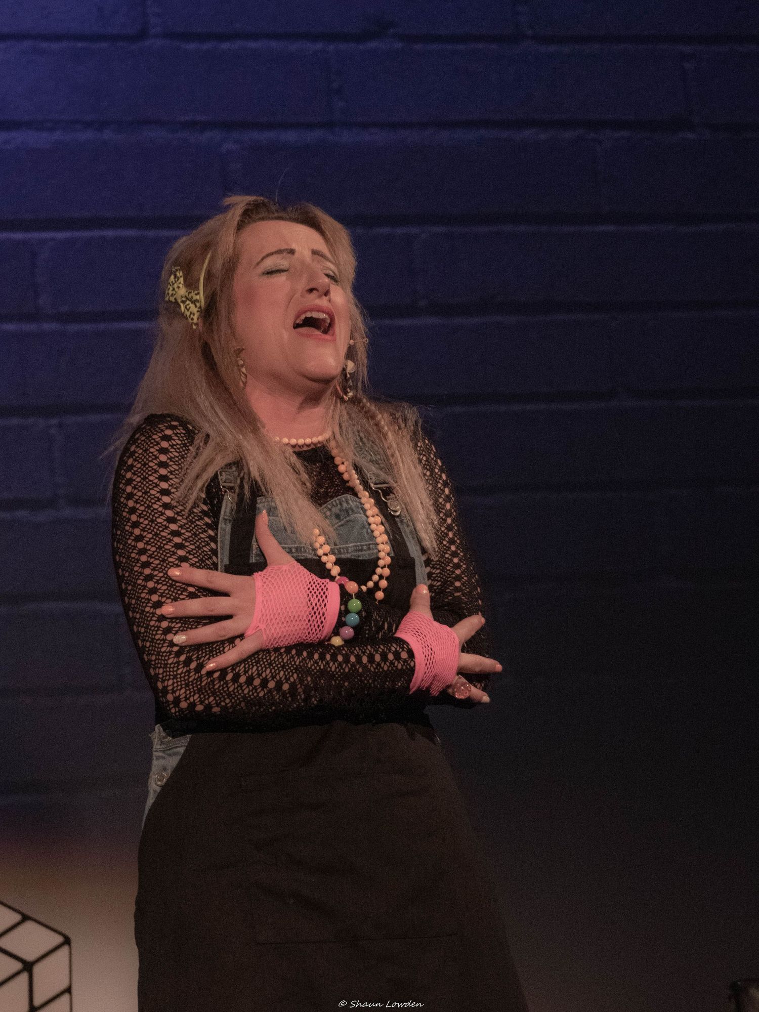 Performer in black mesh top and beaded necklace sings emotionally on stage.