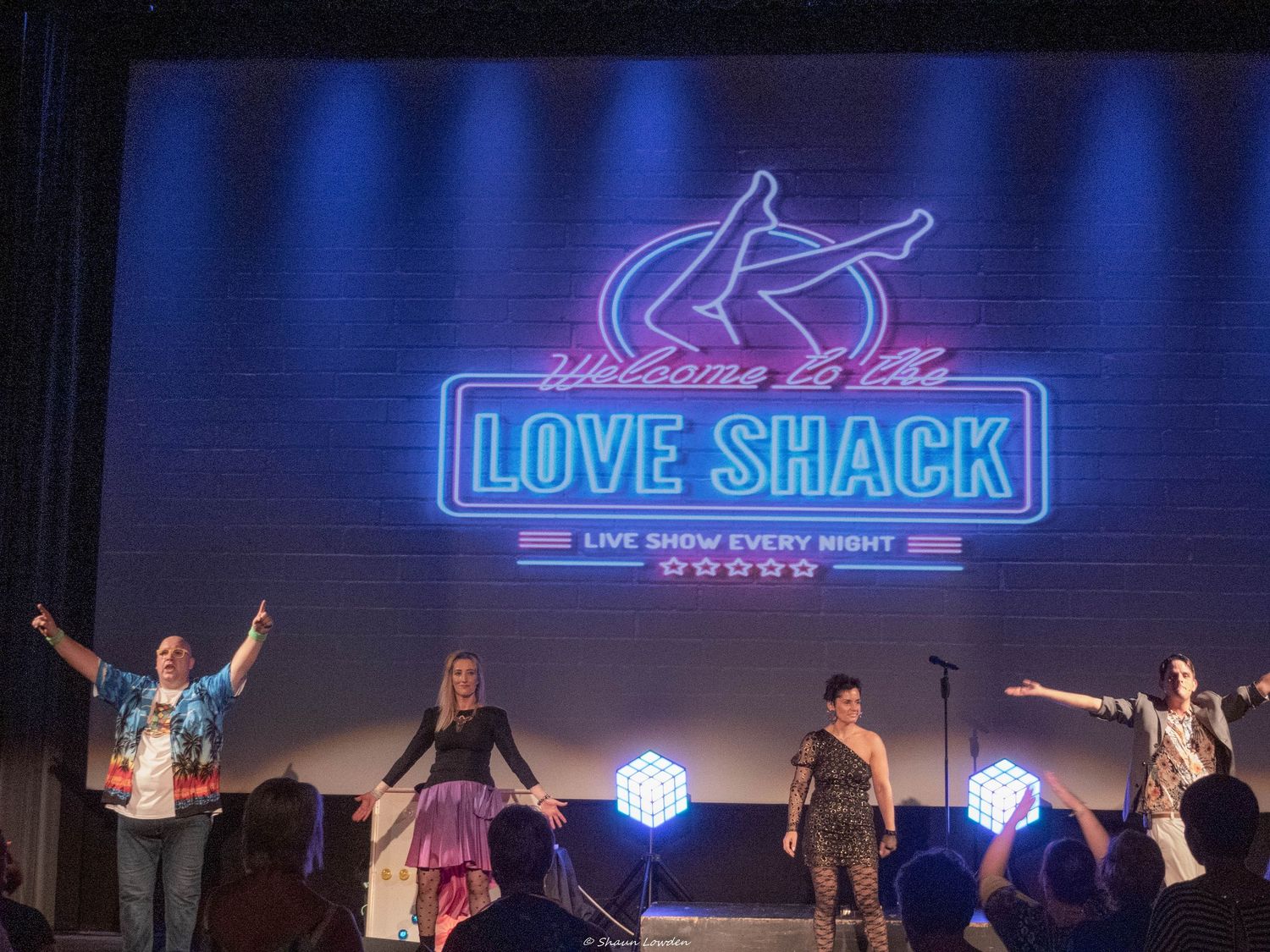 Stage performers pose beneath neon Love Shack sign during theatrical finale.