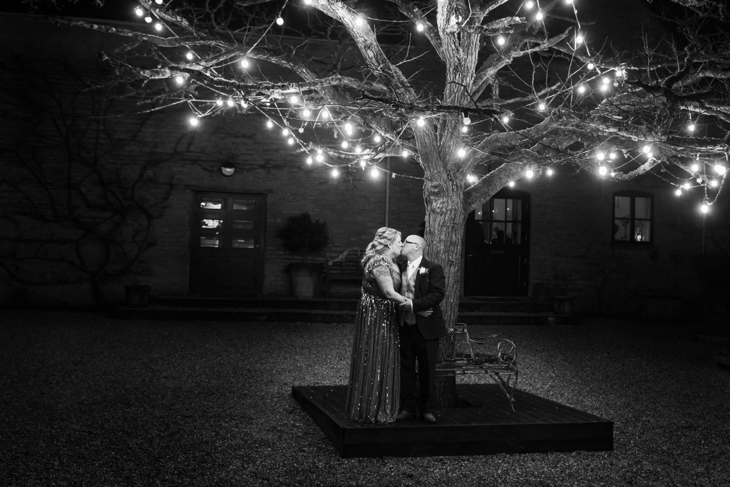 A romantic couple dances beneath a tree decorated with twinkling lights in a nighttime garden setting.