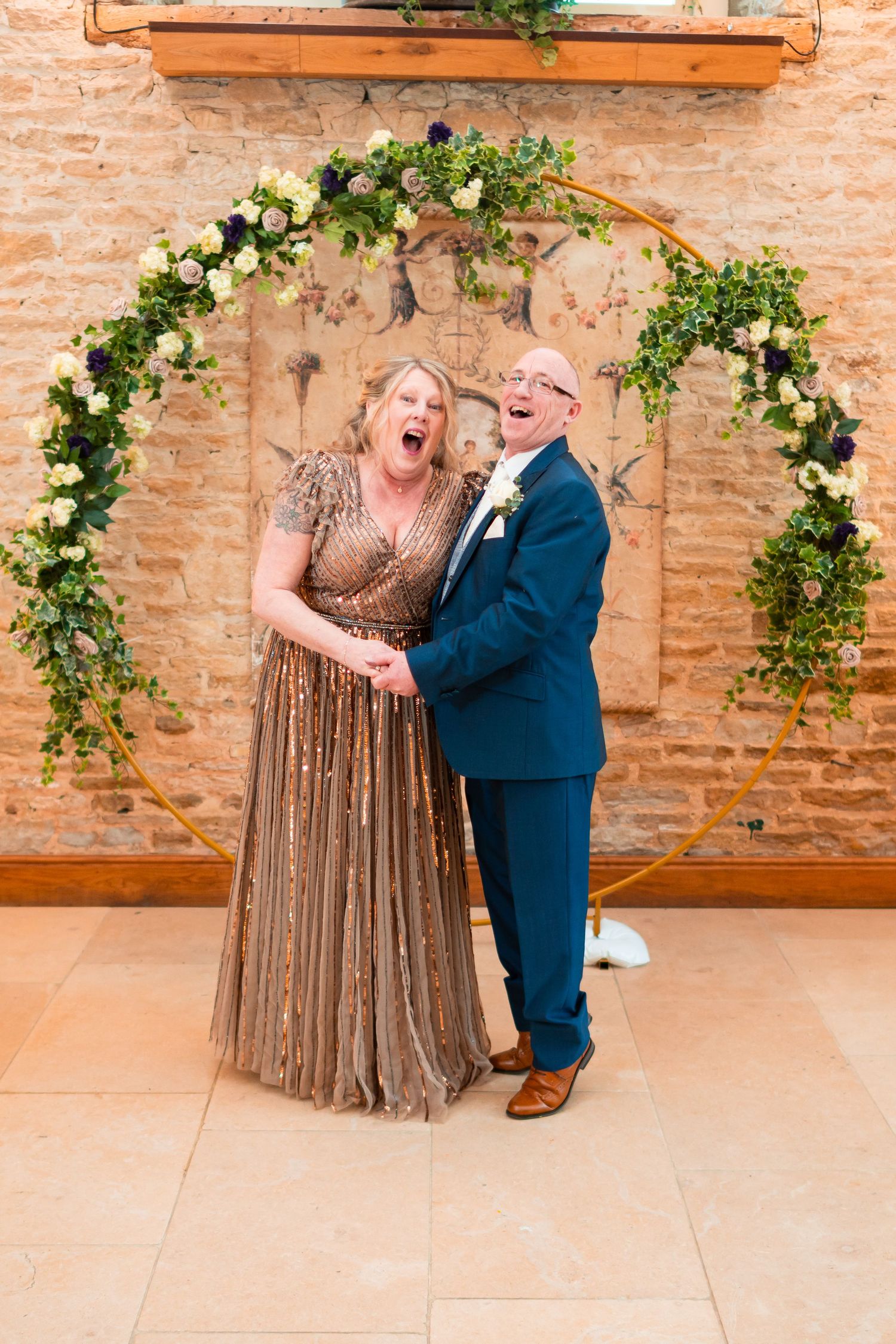 A couple poses joyfully under a floral arch with exposed brick walls in a wedding celebration.