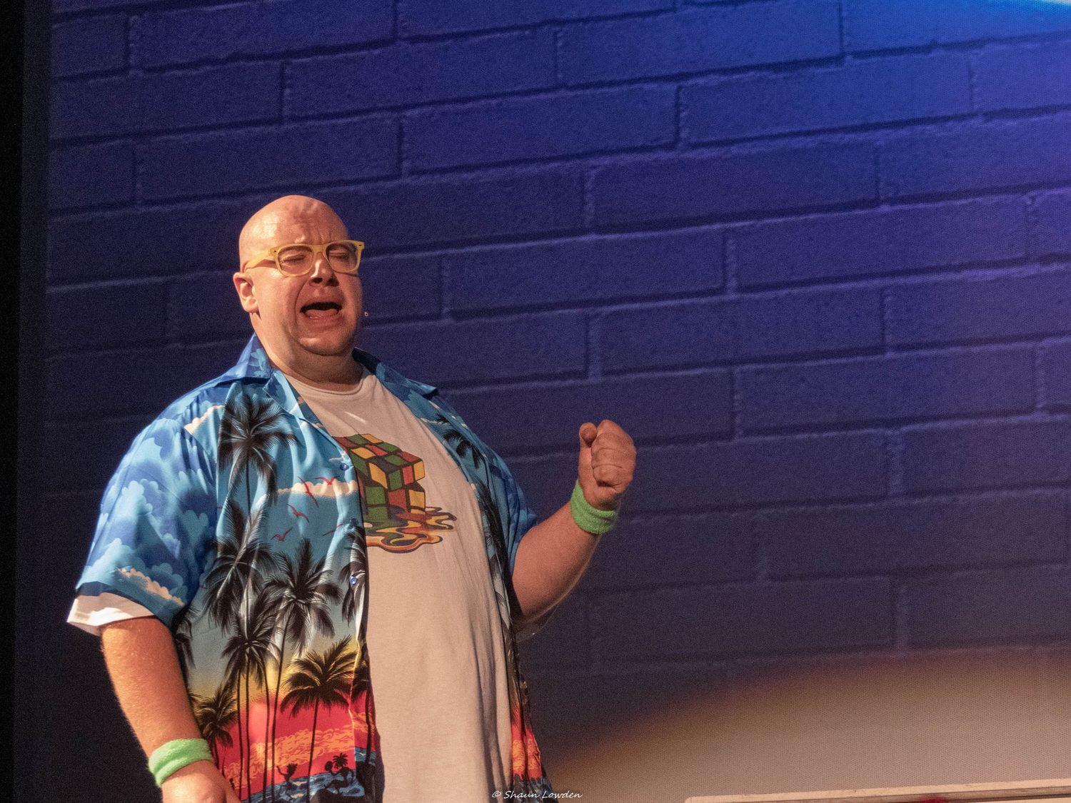 Performer in tropical print shirt gestures expressively against blue wall backdrop.