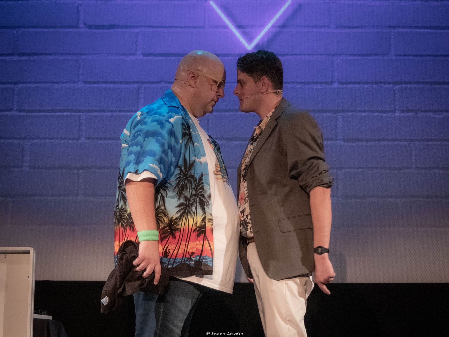 Two performers in conversation during theatrical scene against purple-lit backdrop.