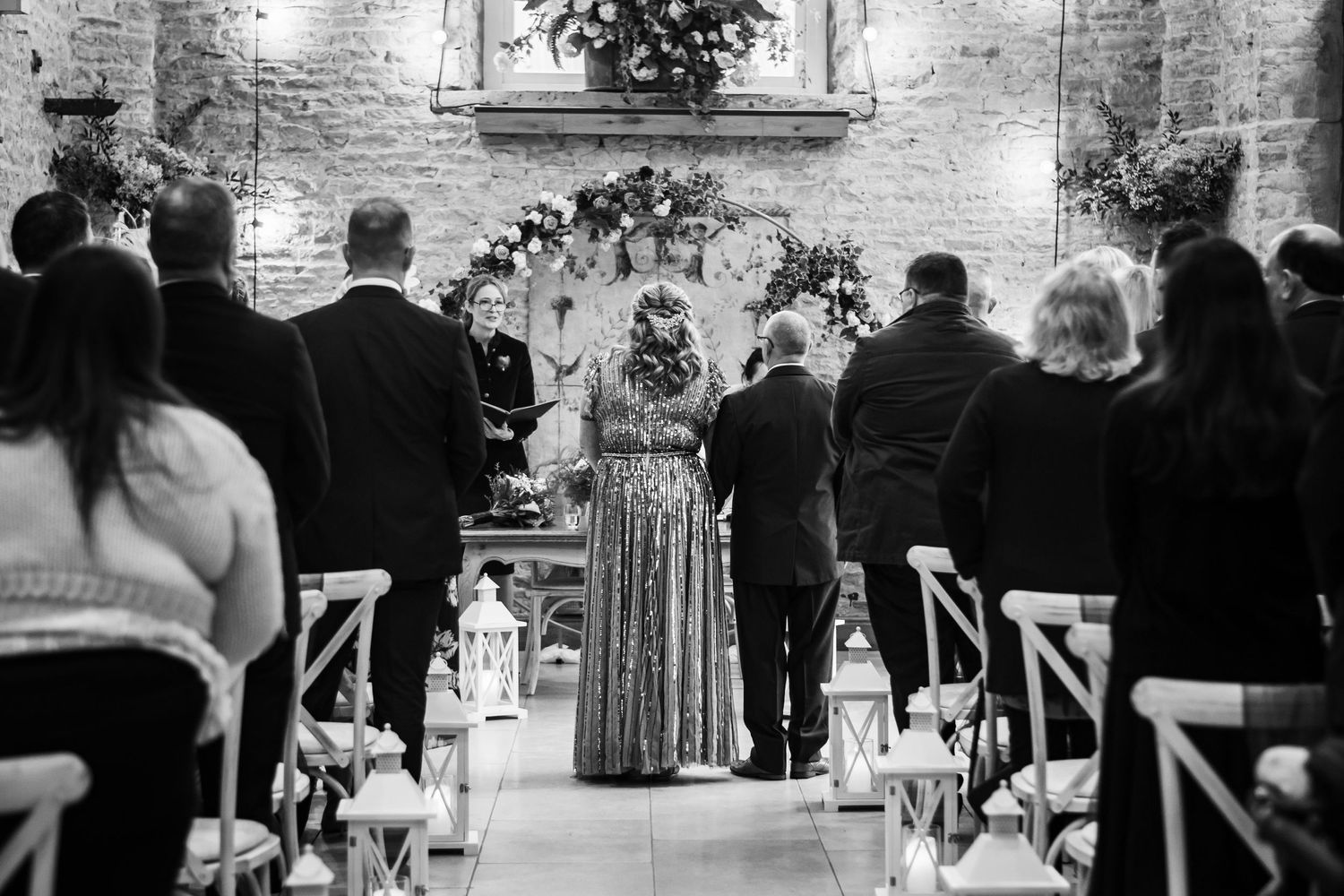 A black and white indoor wedding ceremony scene with guests seated in rows and an officiant conducting the service.