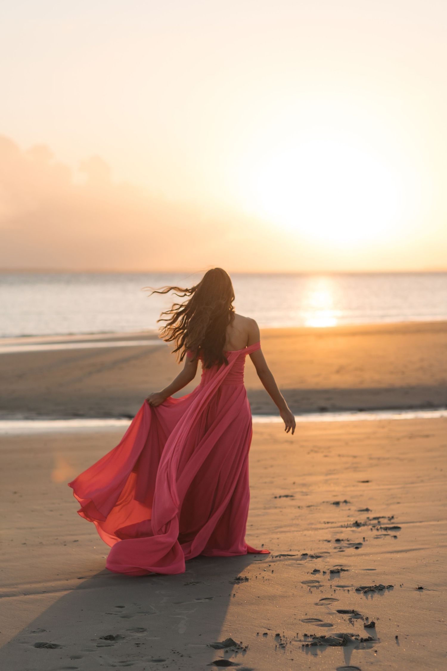 Flowing red dress silhouetted against golden beach sunset with waves in background.