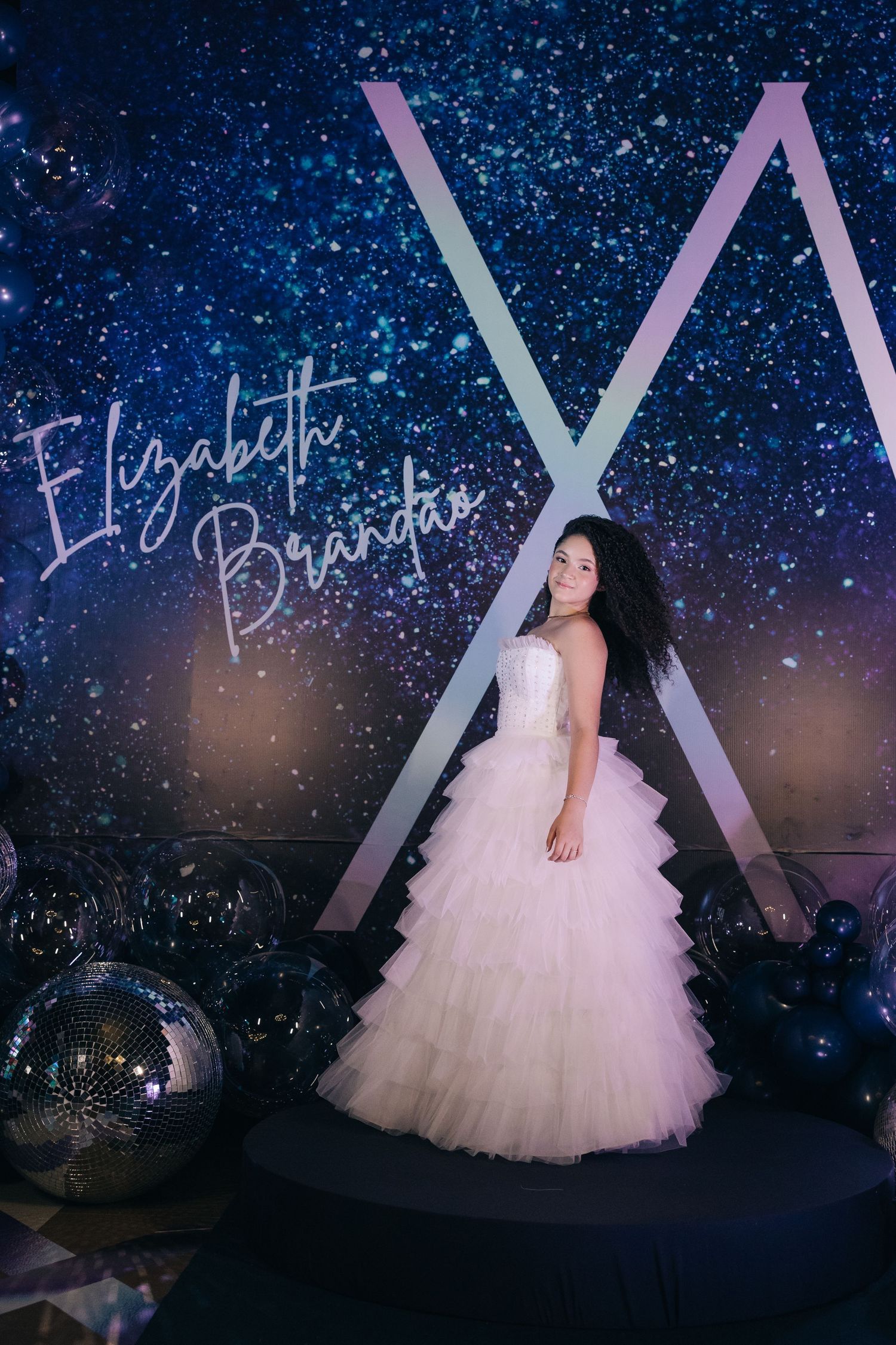 A person in a white layered tulle ball gown poses against a starry night backdrop with geometric lighting elements.