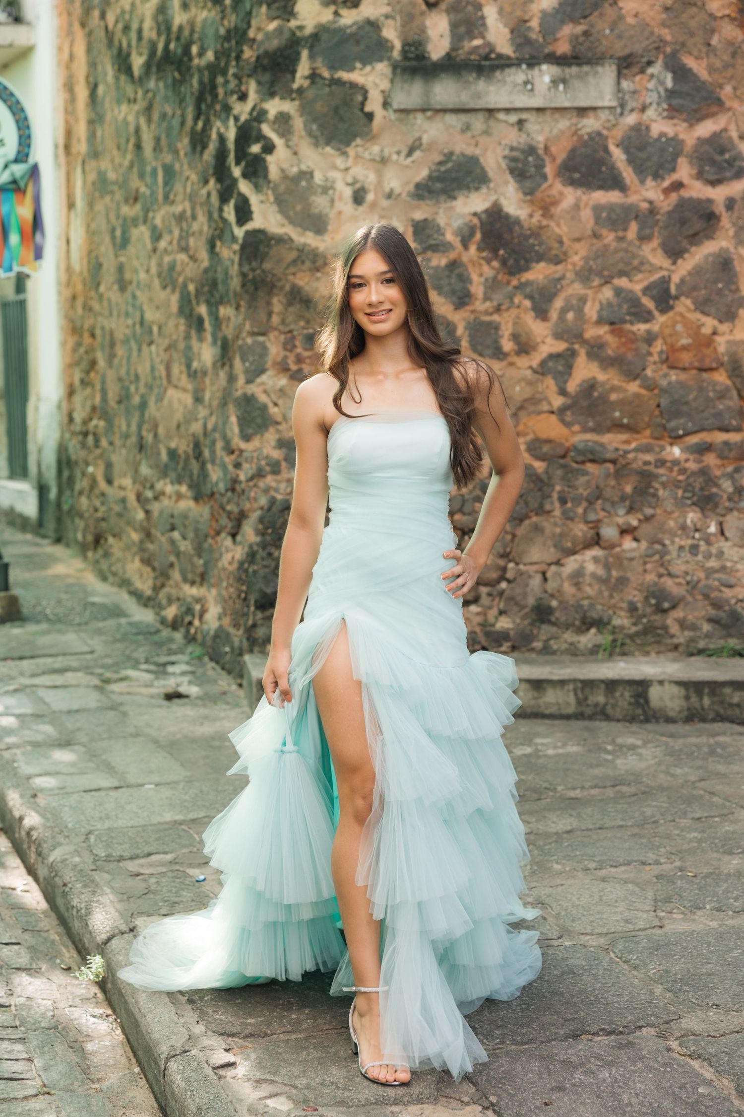 Elegant light blue ruffled gown with high slit photographed against rustic stone wall in urban setting.