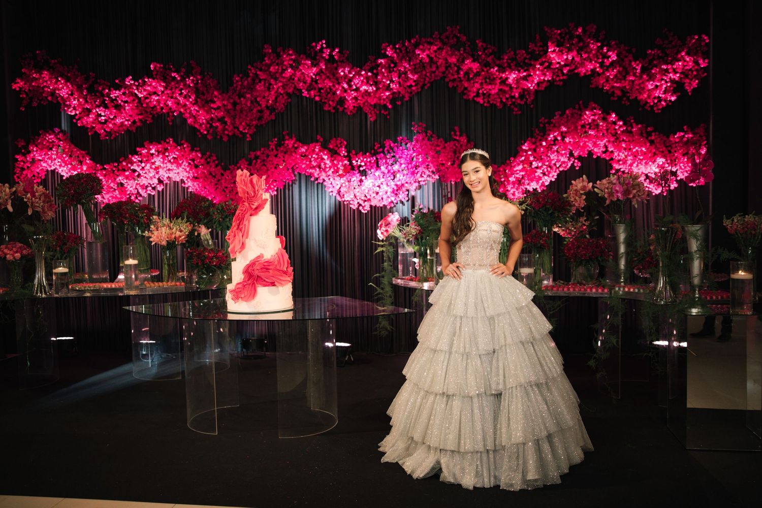 An elegant ballroom decorated with illuminated pink cherry blossom trees and a white tiered ball gown in the center.