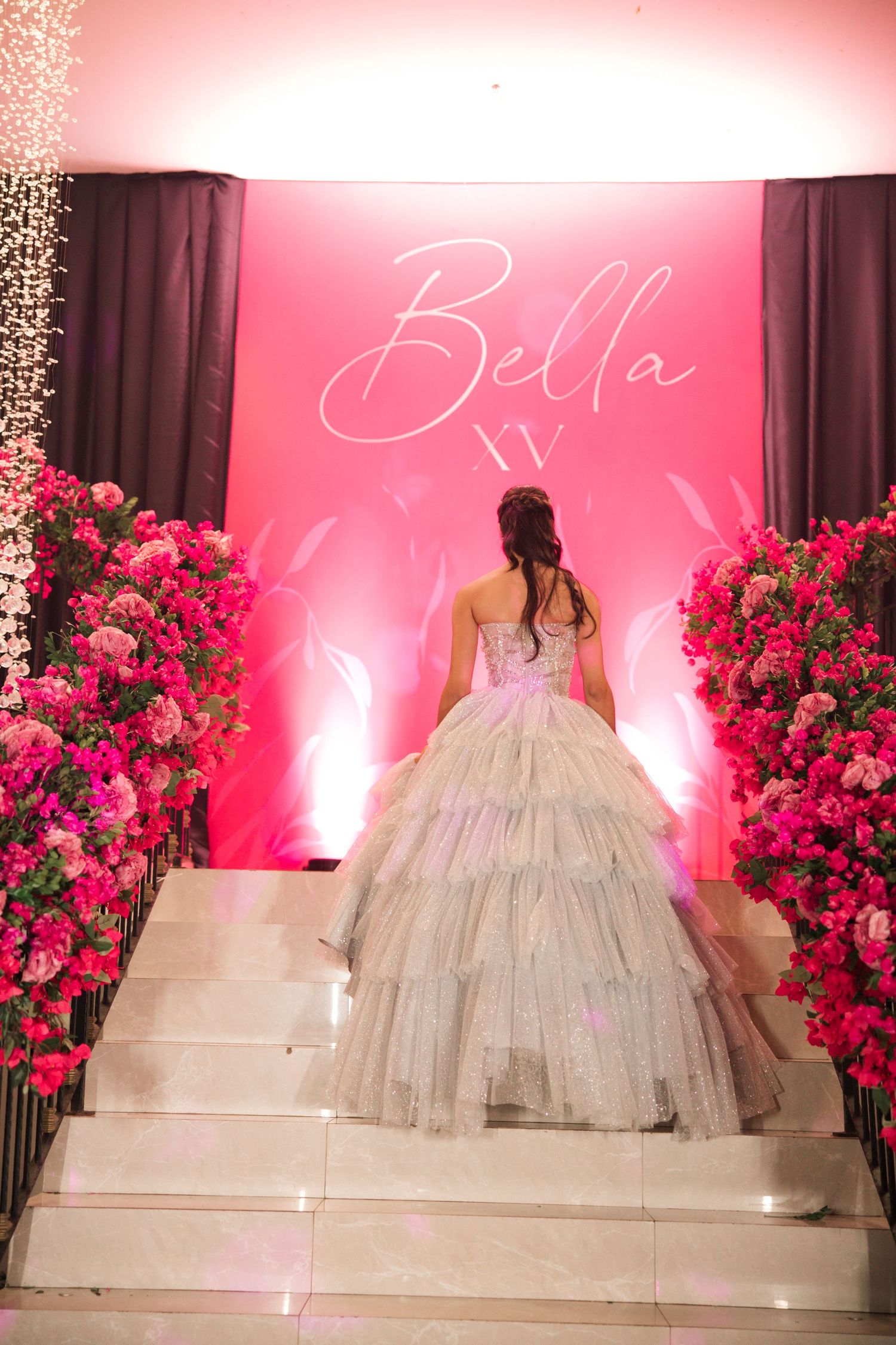 A silver ballgown photographed from behind on white stairs with pink florals and illuminated 'Bella' sign.