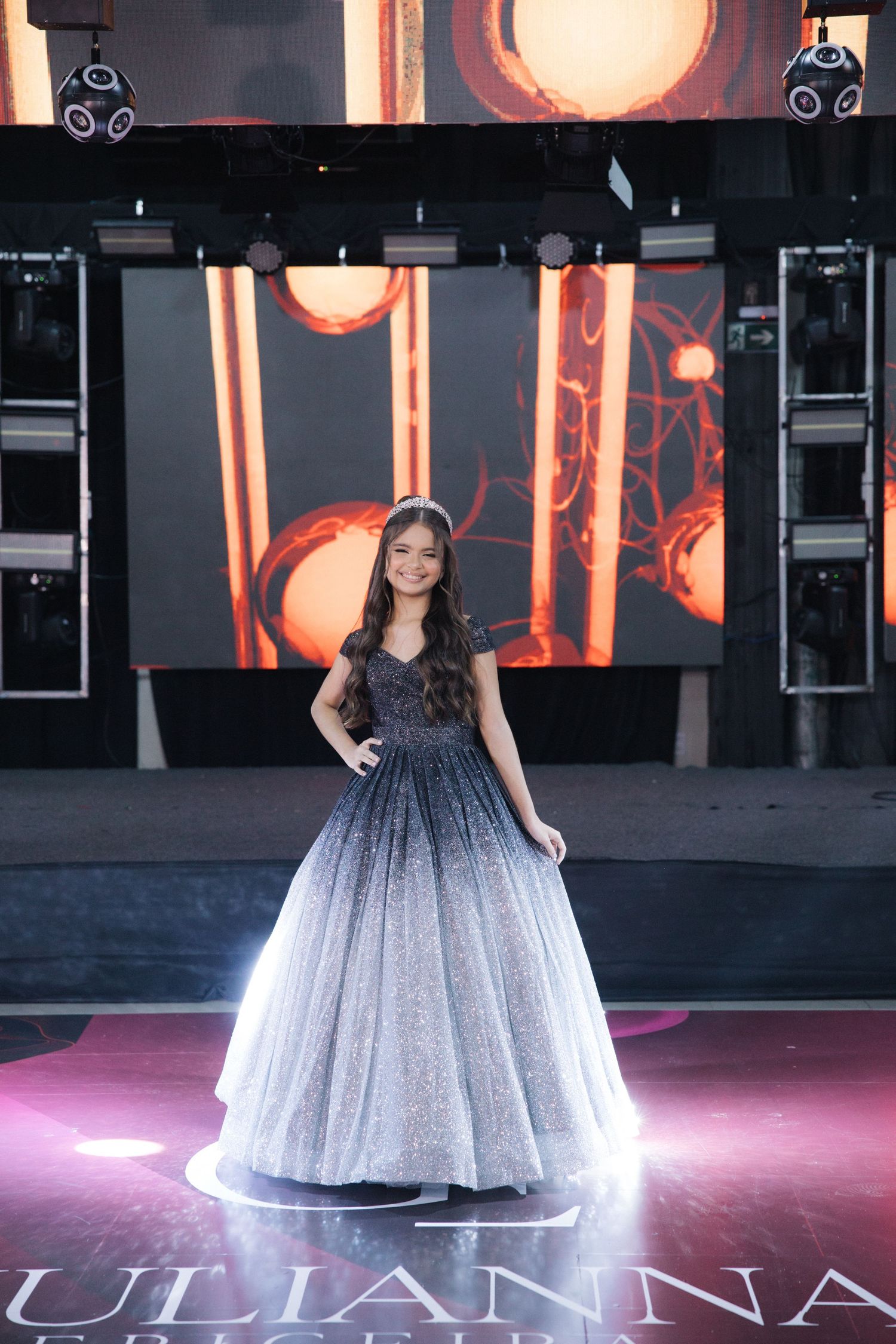 A sparkly ombre ball gown on a dimly lit stage with orange lighting and speakers in the background.