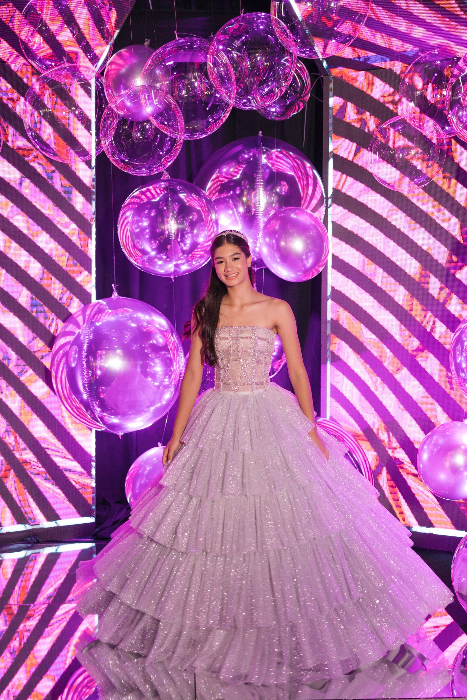 A stunning ballgown dress illuminated by purple lighting and silver balloons creates a magical quinceañera setting.