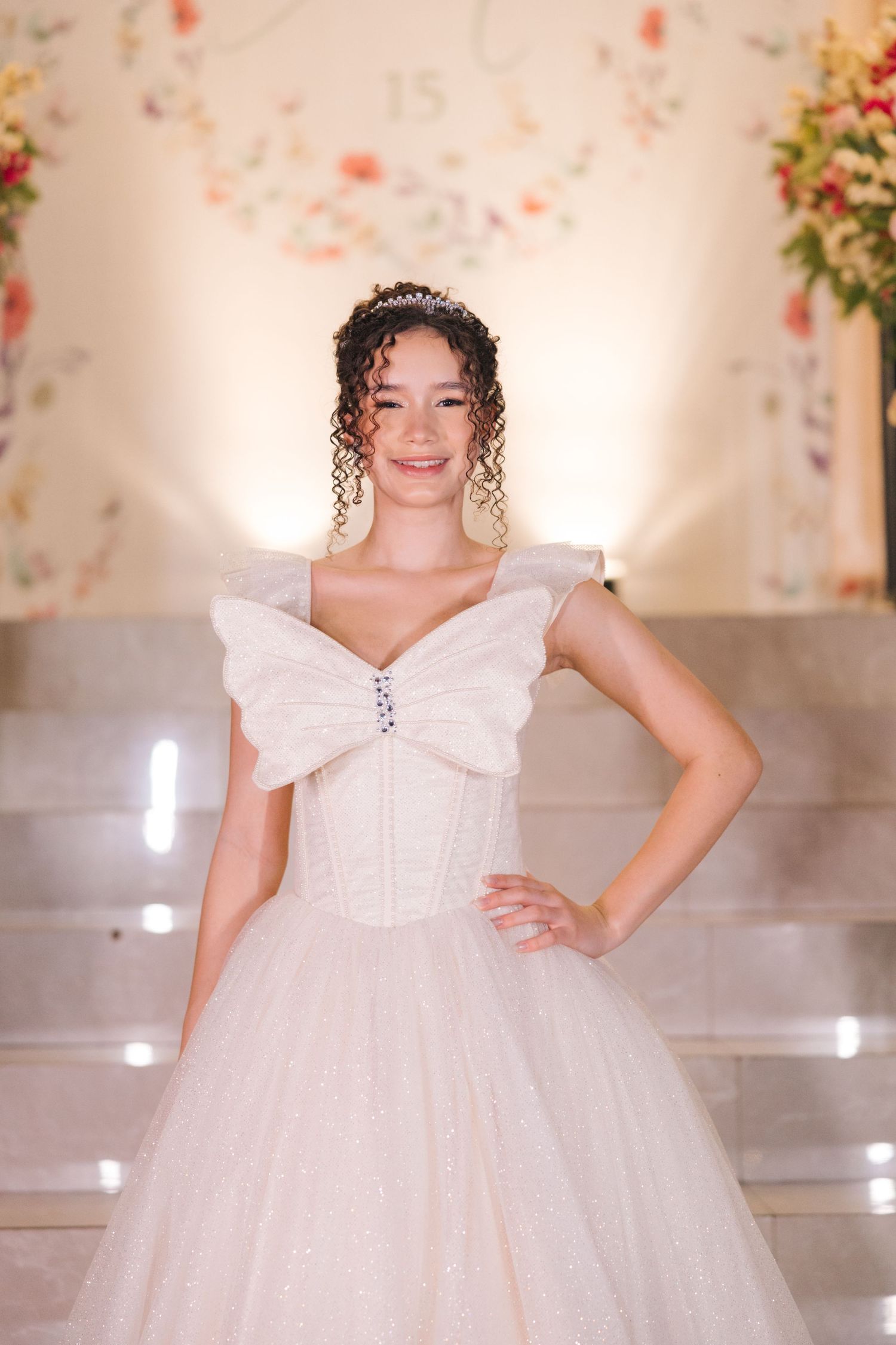 Bride wearing a glamorous white ballgown with bow detail and tiara poses elegantly in front of floral backdrop.