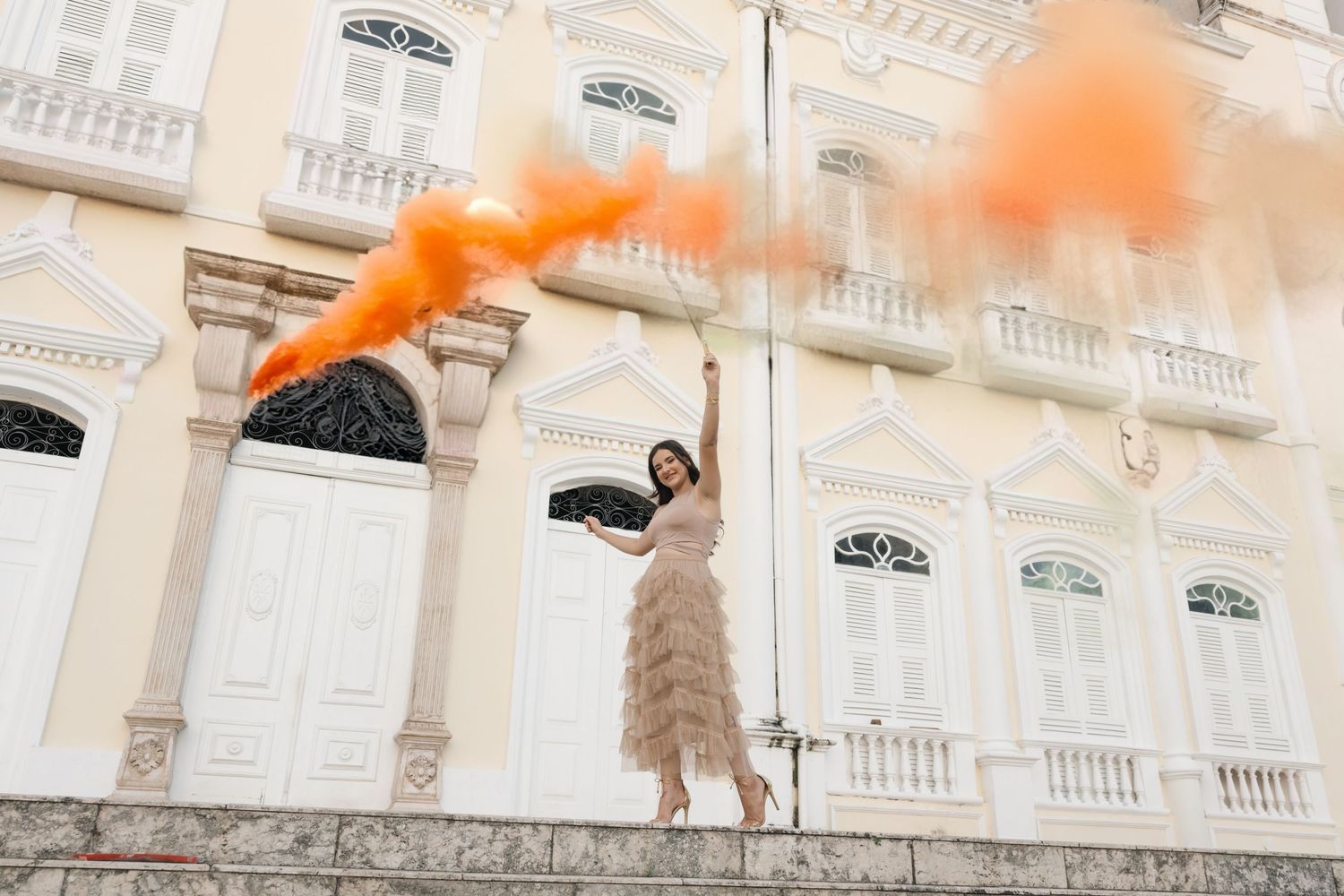 Someone in a flowing dress poses elegantly against classical architecture while orange smoke billows above.