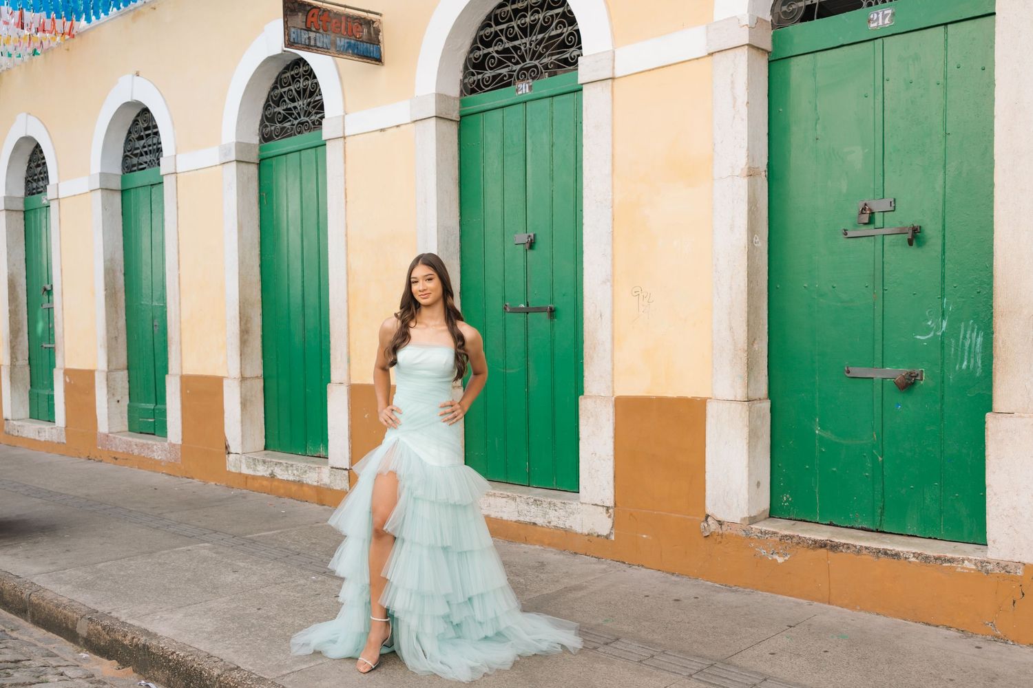 An elegant light blue gown flows along a street with yellow walls and green colonial doors.