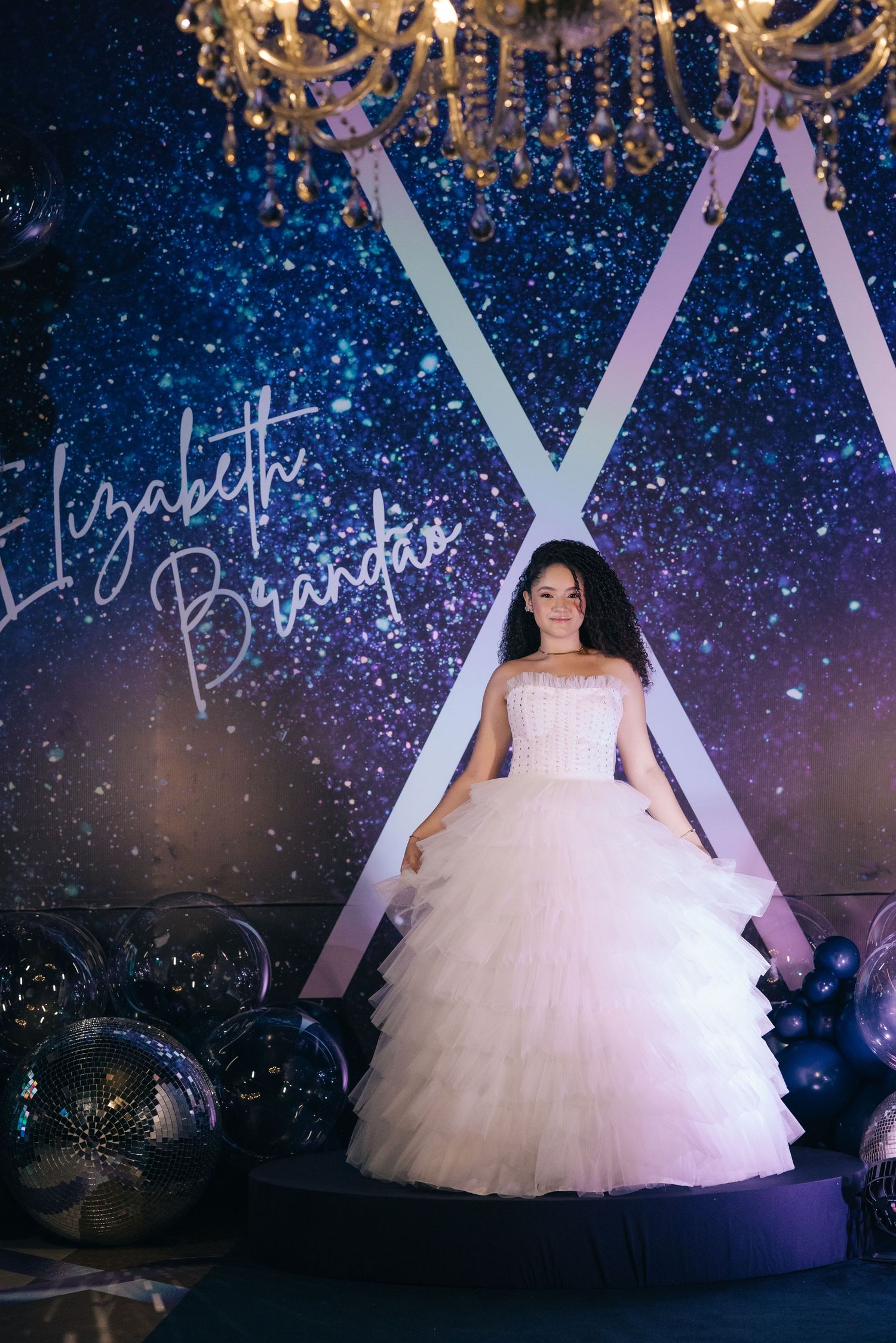 A stunning white ball gown with tiered tulle skirt against a starry backdrop with white draped fabric and chandeliers.
