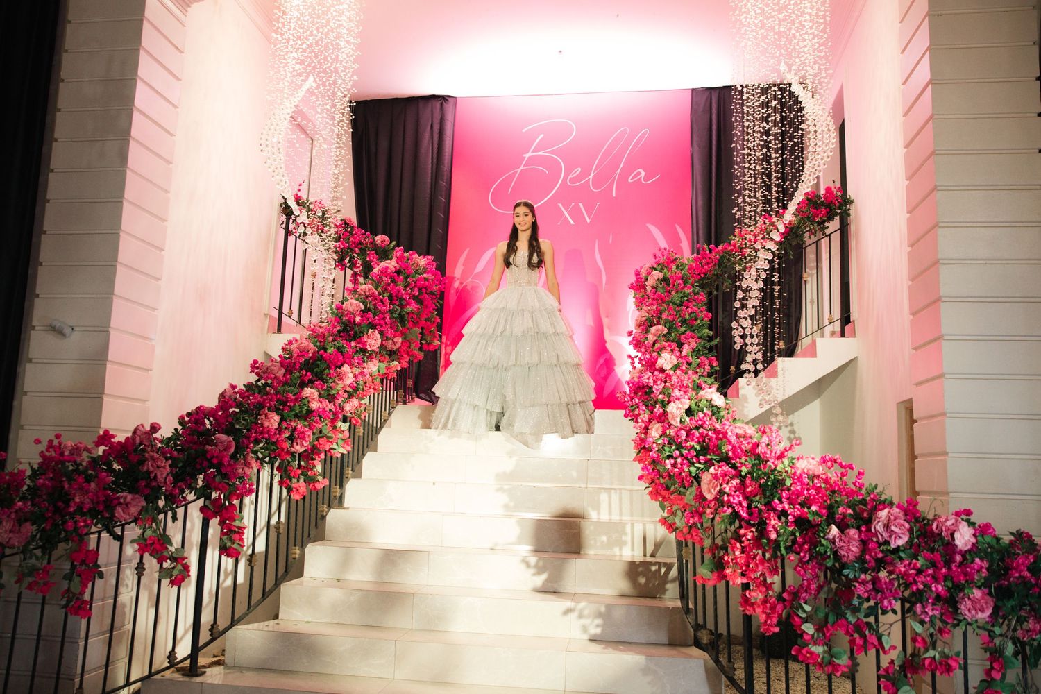White ball gown photographed on grand staircase lined with pink roses and soft lighting.
