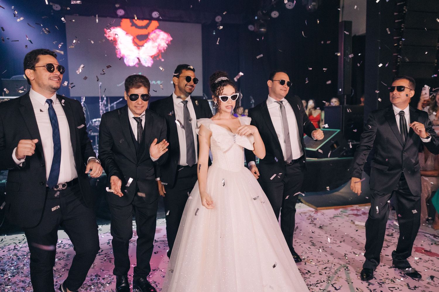 Wedding party in suits and sunglasses dancing in formation on confetti-covered floor.