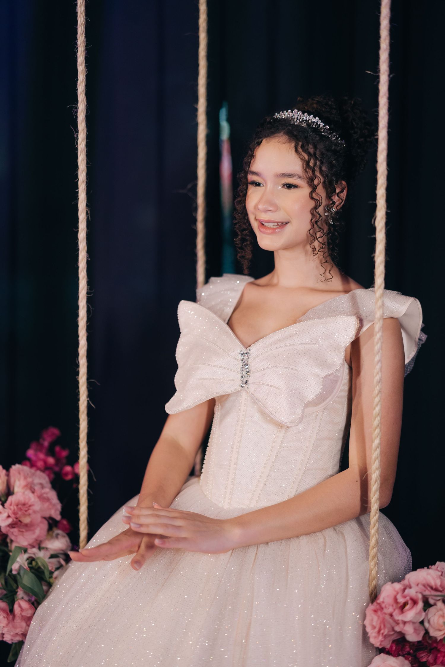 A woman in a sparkly white princess gown and tiara sits elegantly on a swing decorated with pink flowers.