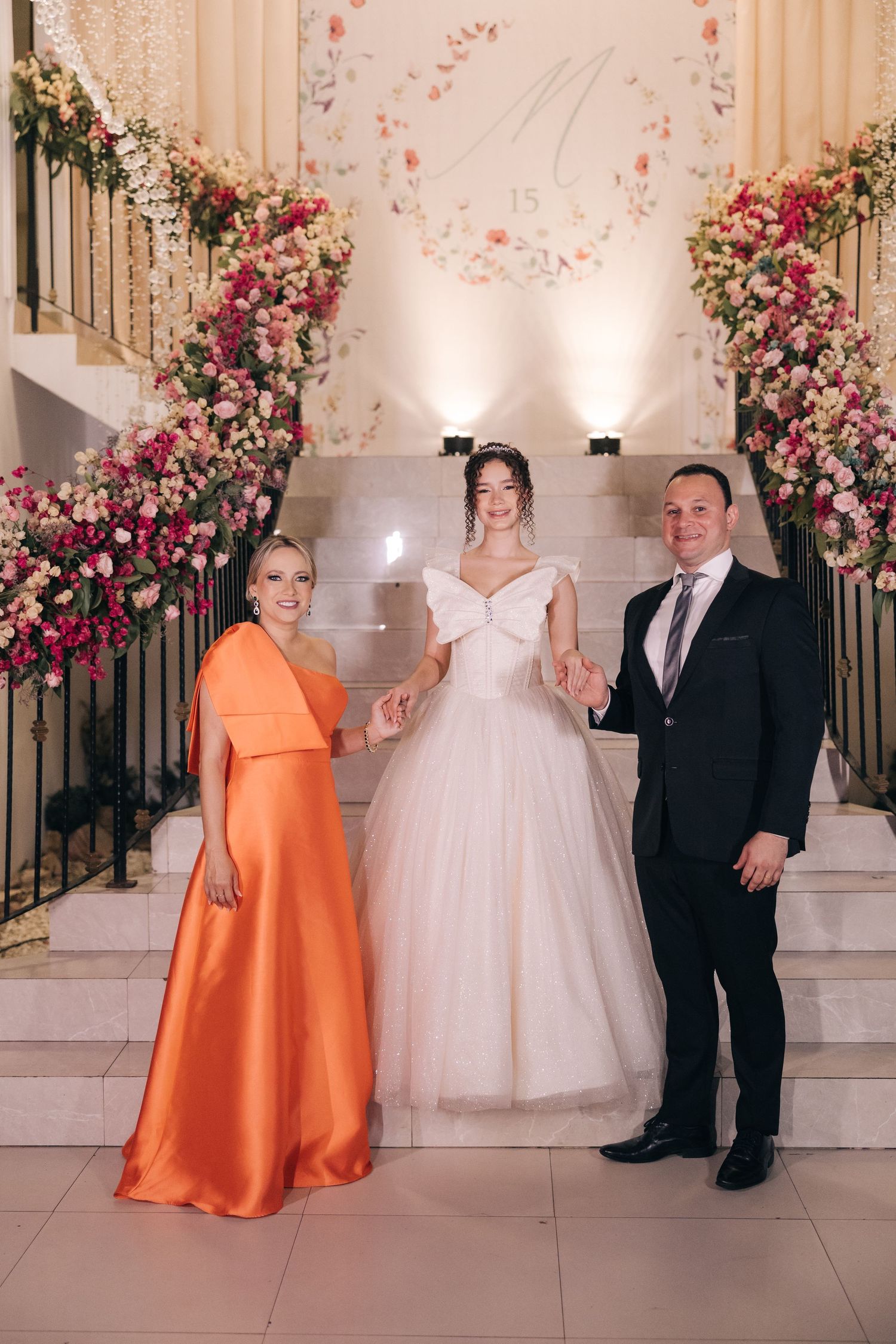 A bride in a white ballgown stands on stairs decorated with pink floral arrangements alongside others in formal attire.