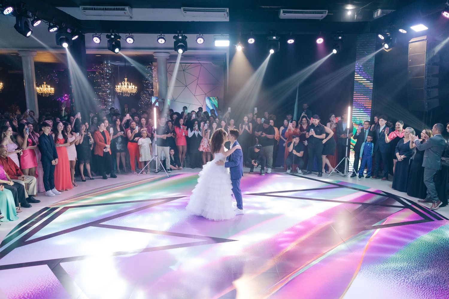 A couple performs their first dance on a lit-up dance floor surrounded by wedding guests in an elegant venue.