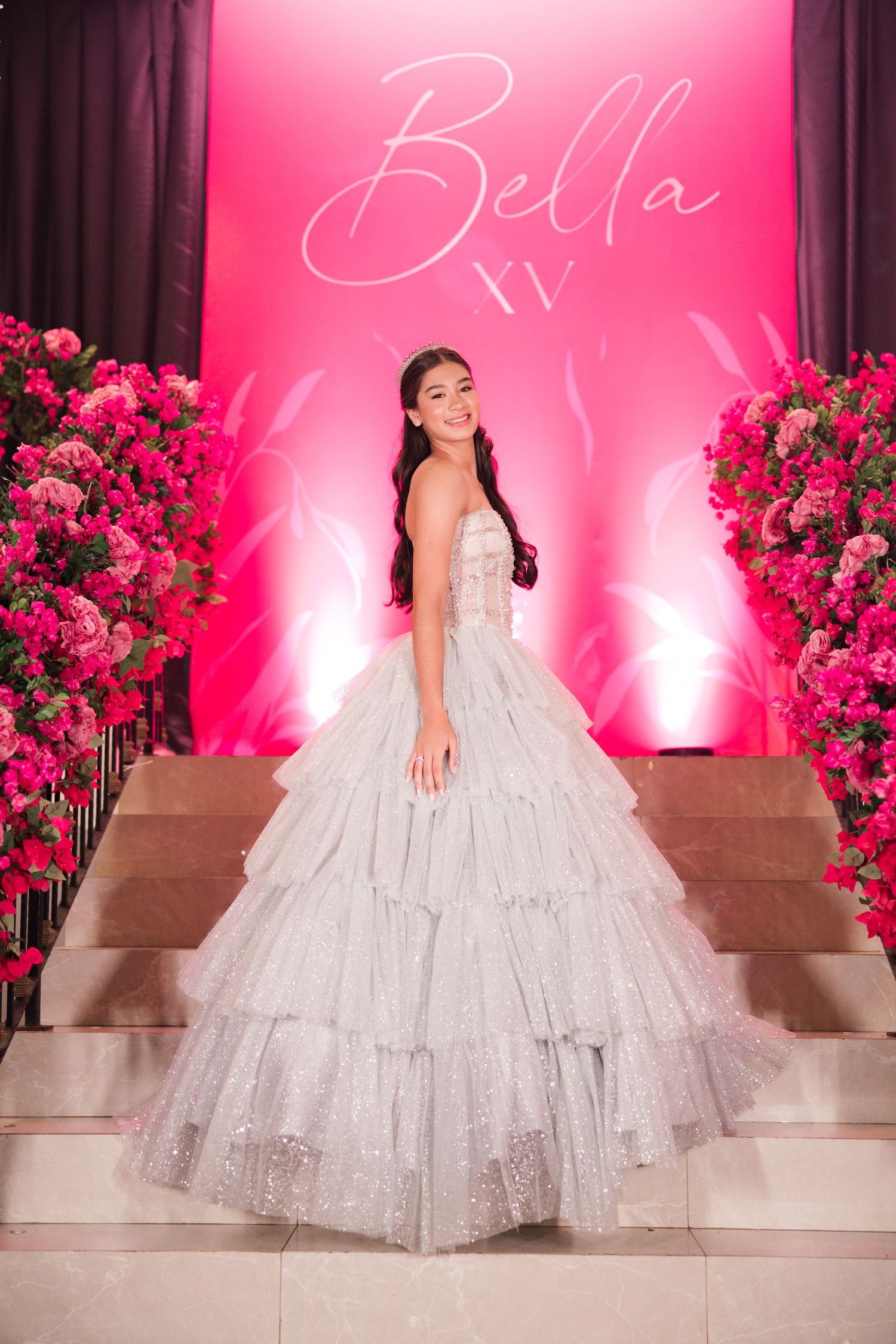 Glamorous silver quinceañera dress photographed against pink backdrop with rose arrangements.