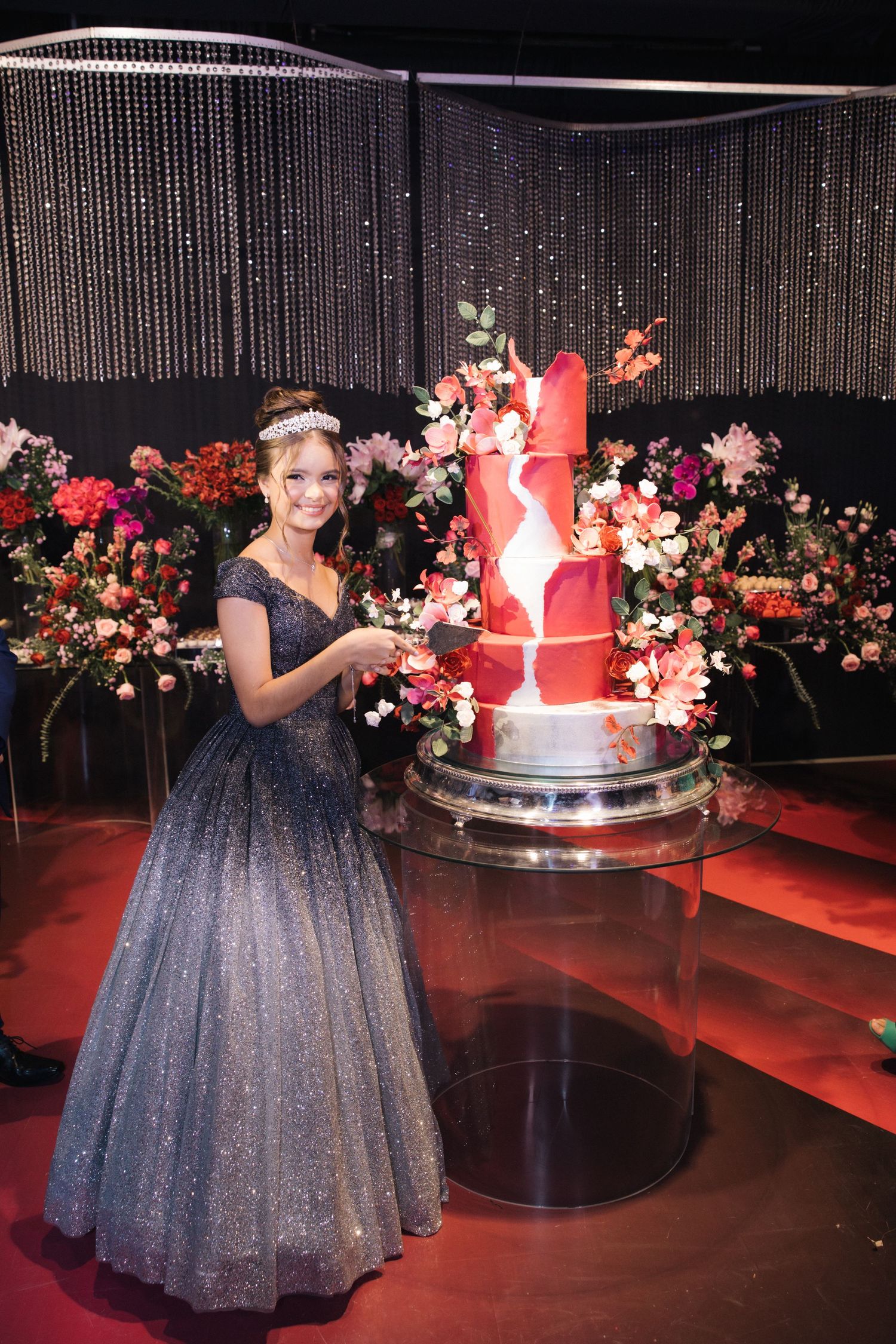 A princess ball gown in glittering ombre stands beside a tall red cake decorated with flowers.