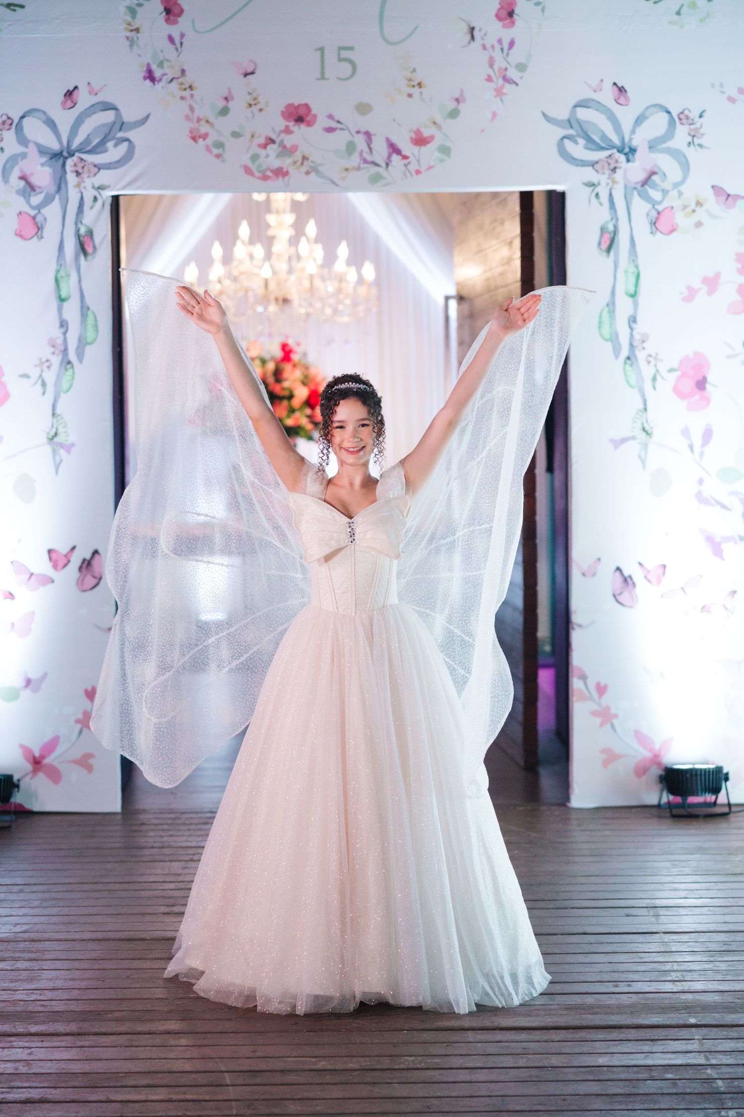 A bride in a flowing white tulle gown raises her veil dramatically while standing in an elegant floral-decorated venue.