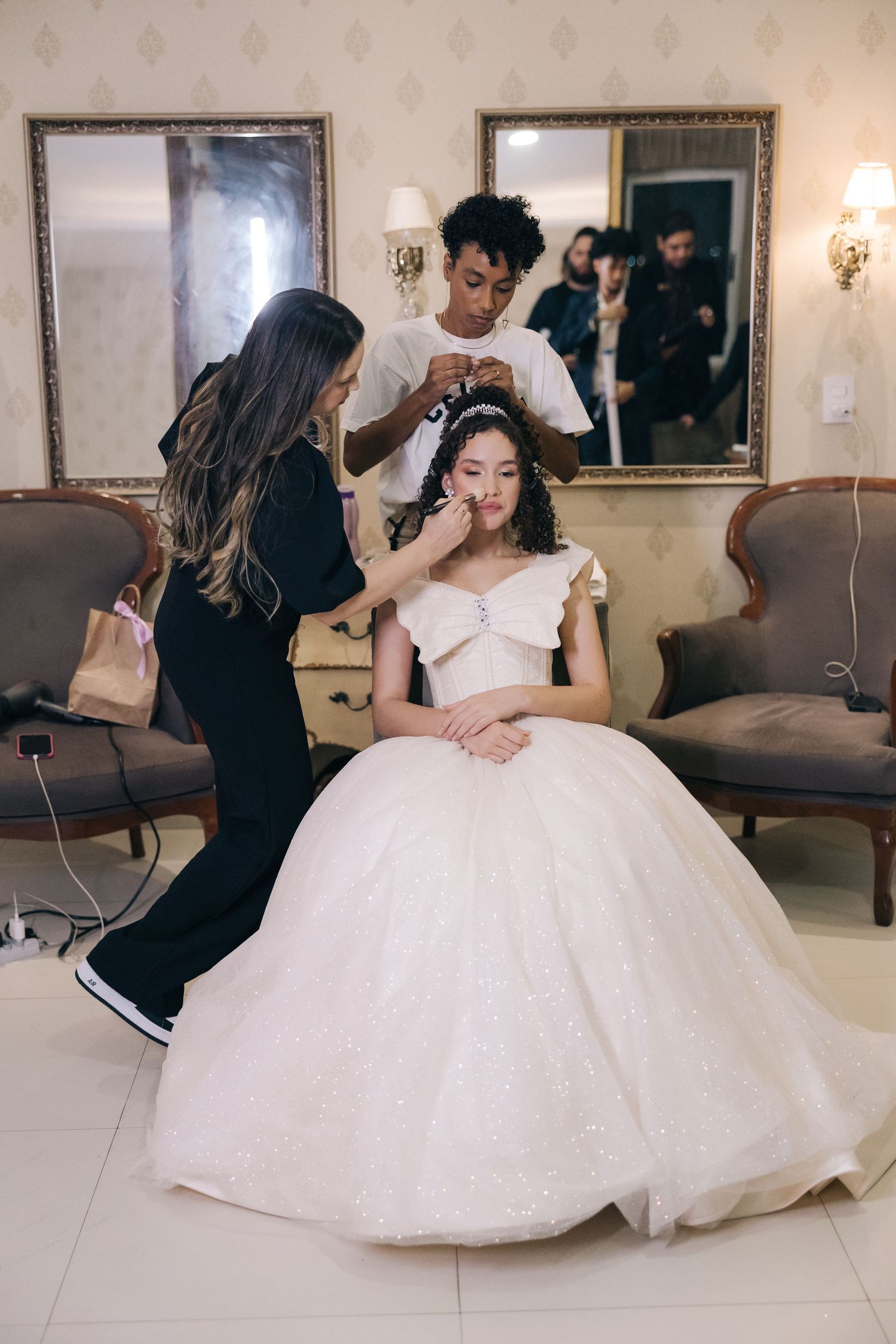 Wedding preparation scene with stylists working on bride's hair and makeup in an elegant room.