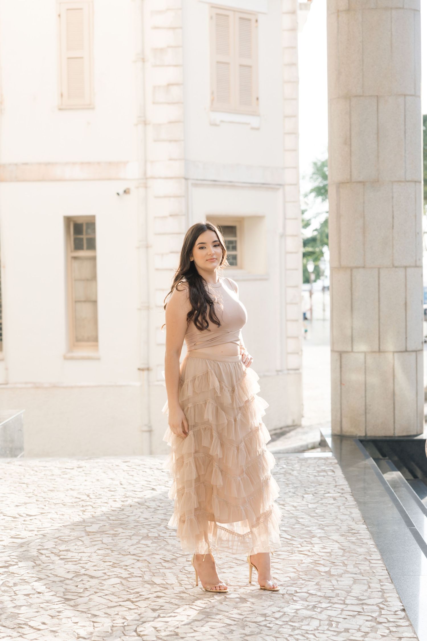A person wearing a blush-colored ruffled maxi dress stands elegantly in front of a light-colored stone building.